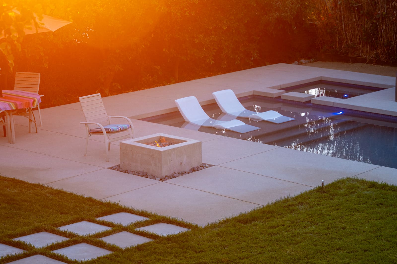 A finished Southern California poolside outdoor living space at sunset with a concrete fire pit, lounge chairs and a spa