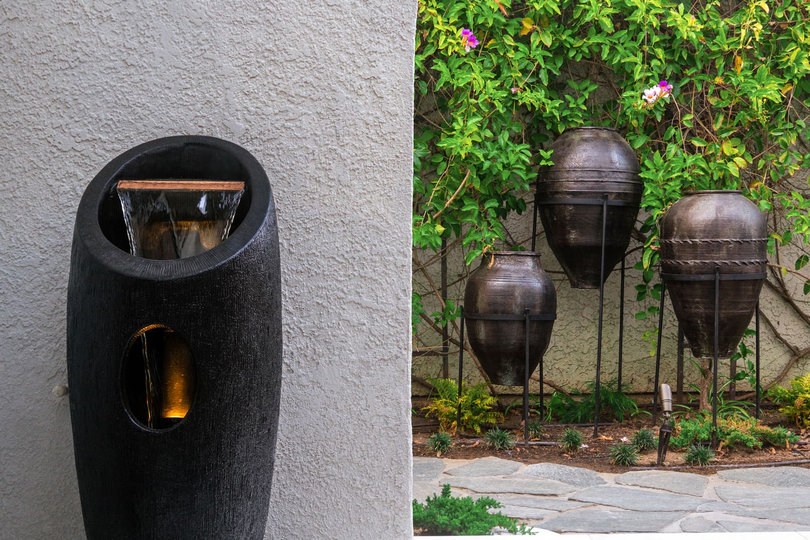 Dark vessel fountain against the stucco wall, with the hanging urns behind