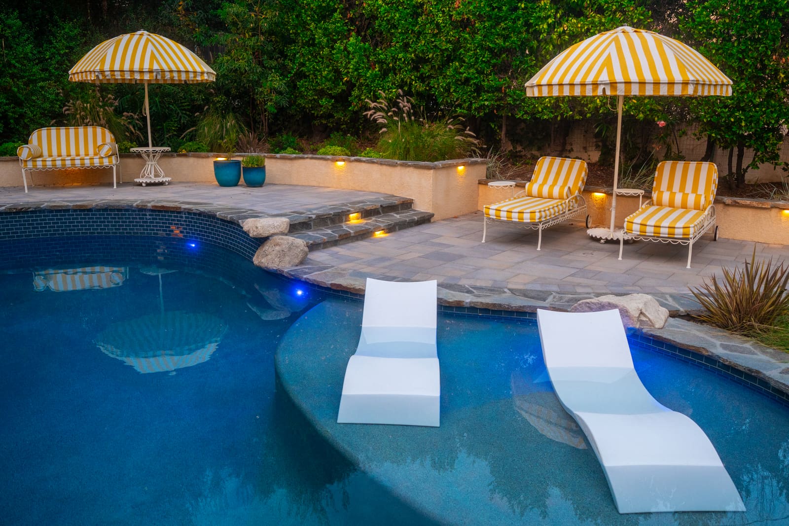The pool at dusk, lit from within, with in-pool loungers and a flagstone deck