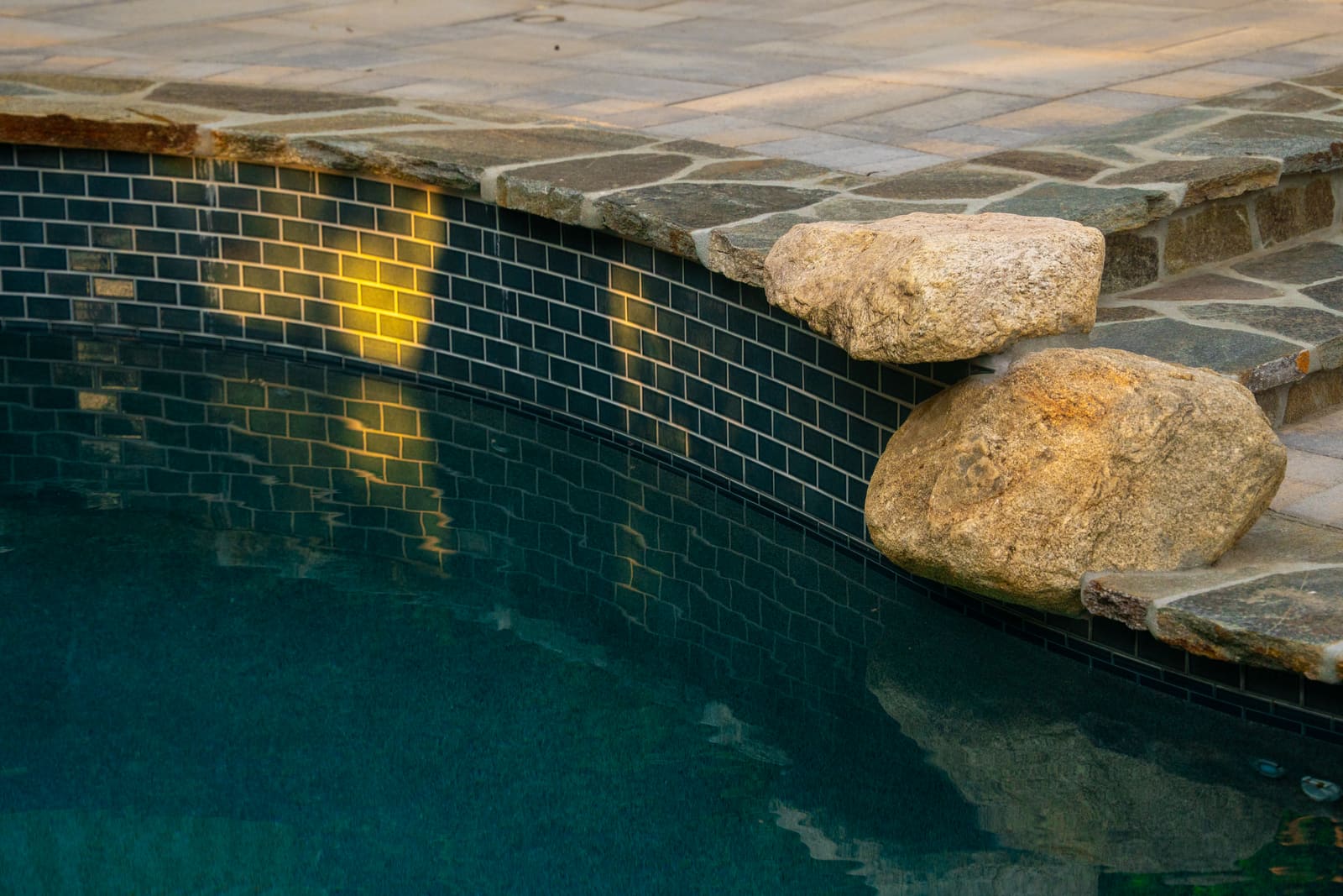 Blue subway-tiled pool wall meeting a granite boulder set into the coping