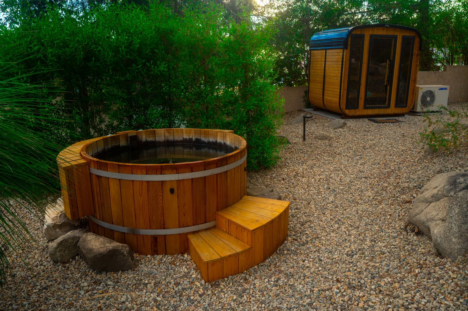 Cedar barrel hot tub with timber steps on a gravel terrace, beside a cedar sauna