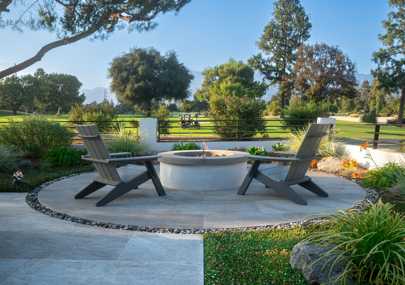Wooden Adirondack chairs around the concrete fire pit, with the golf course fairway and mature trees beyond