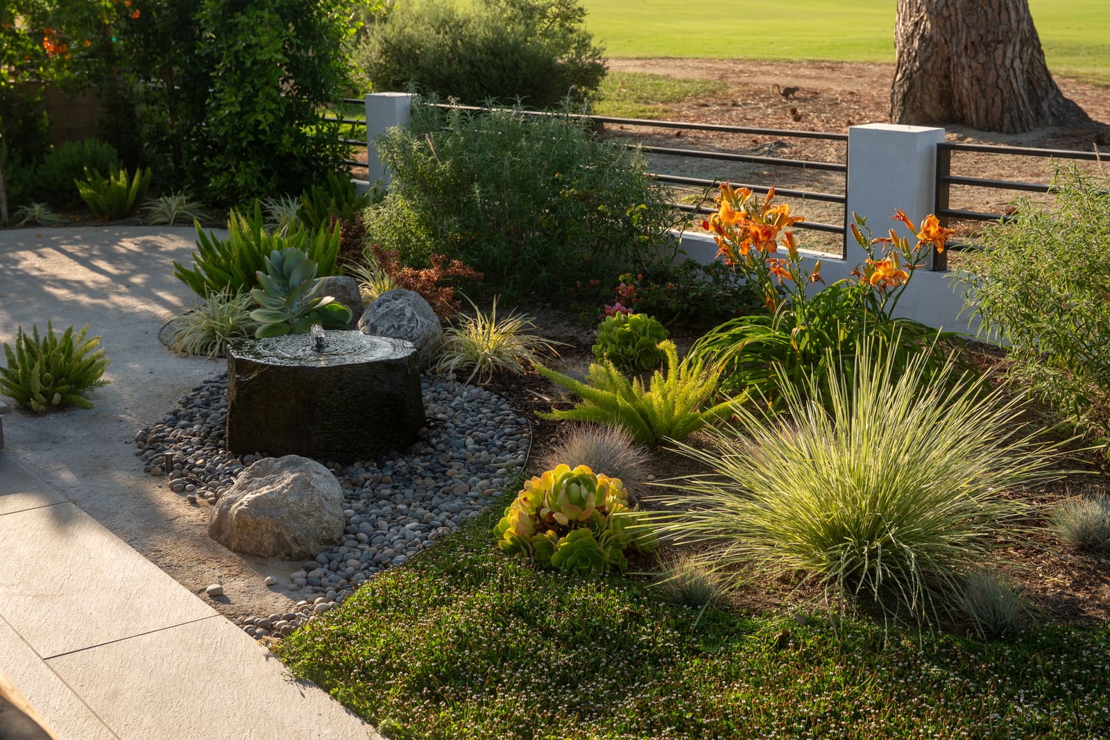 Core-drilled stone column fountain set in a river rock bed, among succulents and grasses
