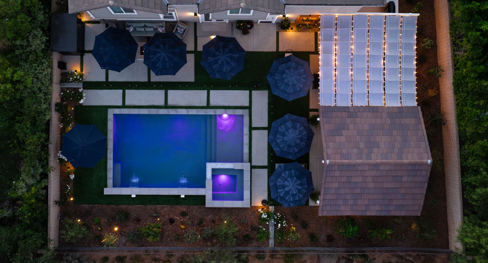 Aerial view at night of the finished yard, with the lit pool, umbrella-shaded dining terrace, cabana and fire table