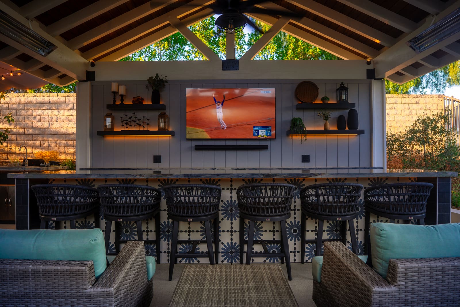Under the cabana's timber roof, a shiplap wall carries a screen, open shelves and lantern lighting above the bar