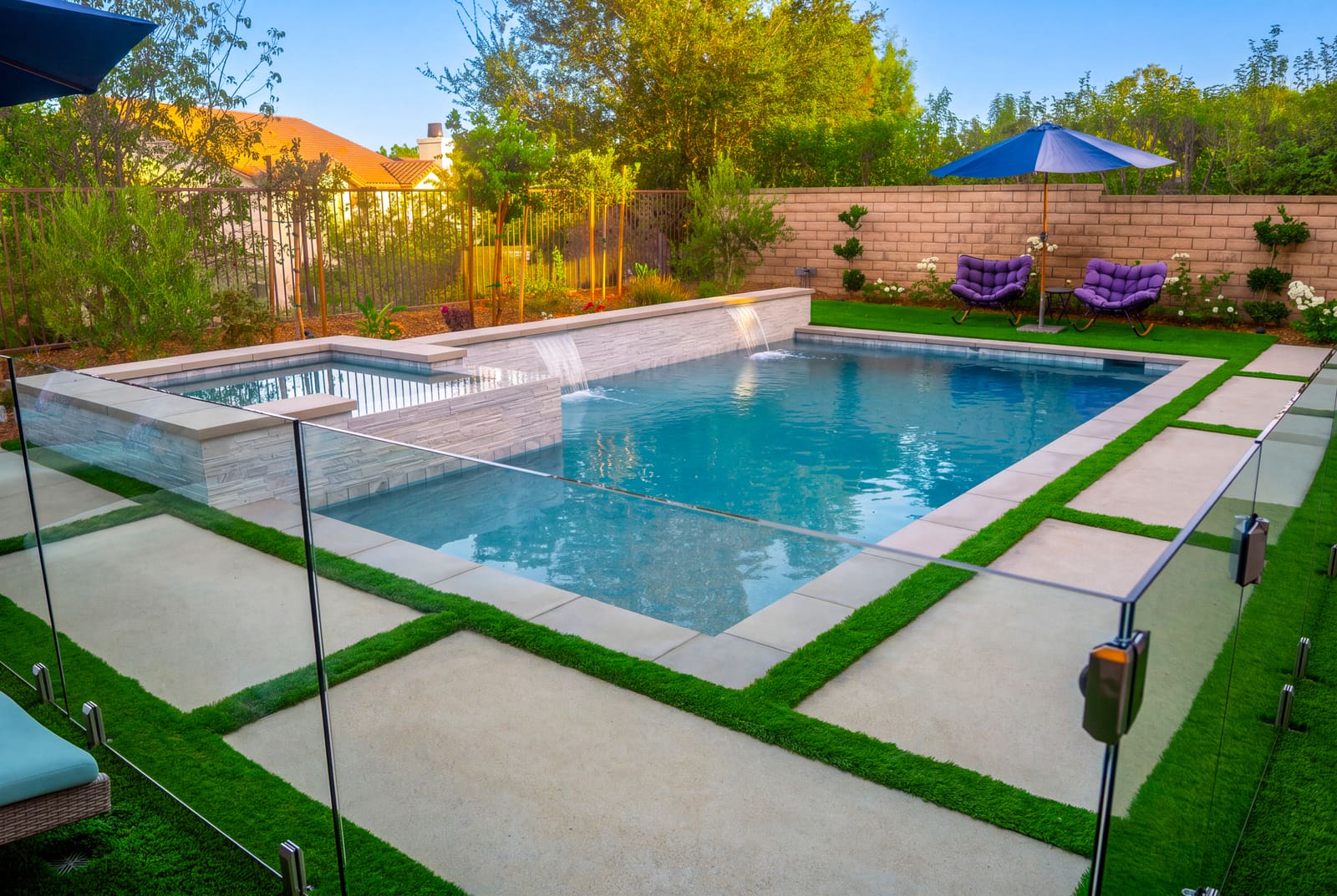 Rectangular pool and raised spa in a field of concrete pads with artificial turf joints, behind a glass safety fence