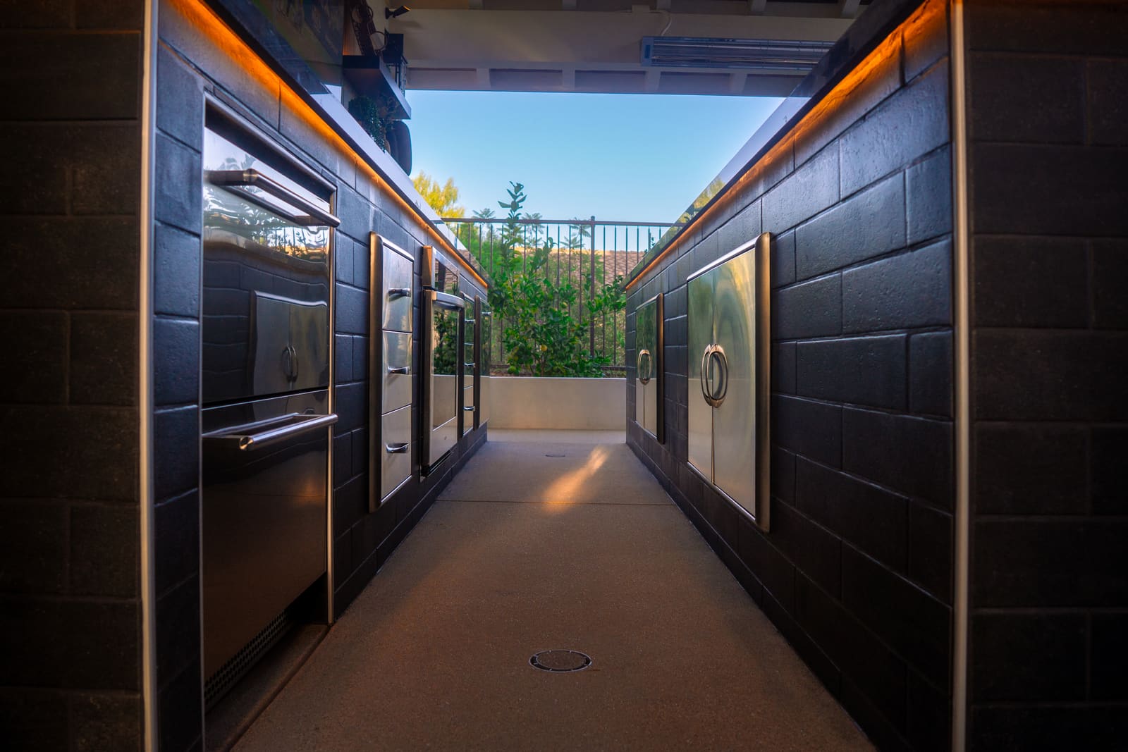The outdoor kitchen at dusk, stainless appliances lit along both dark cabinet runs