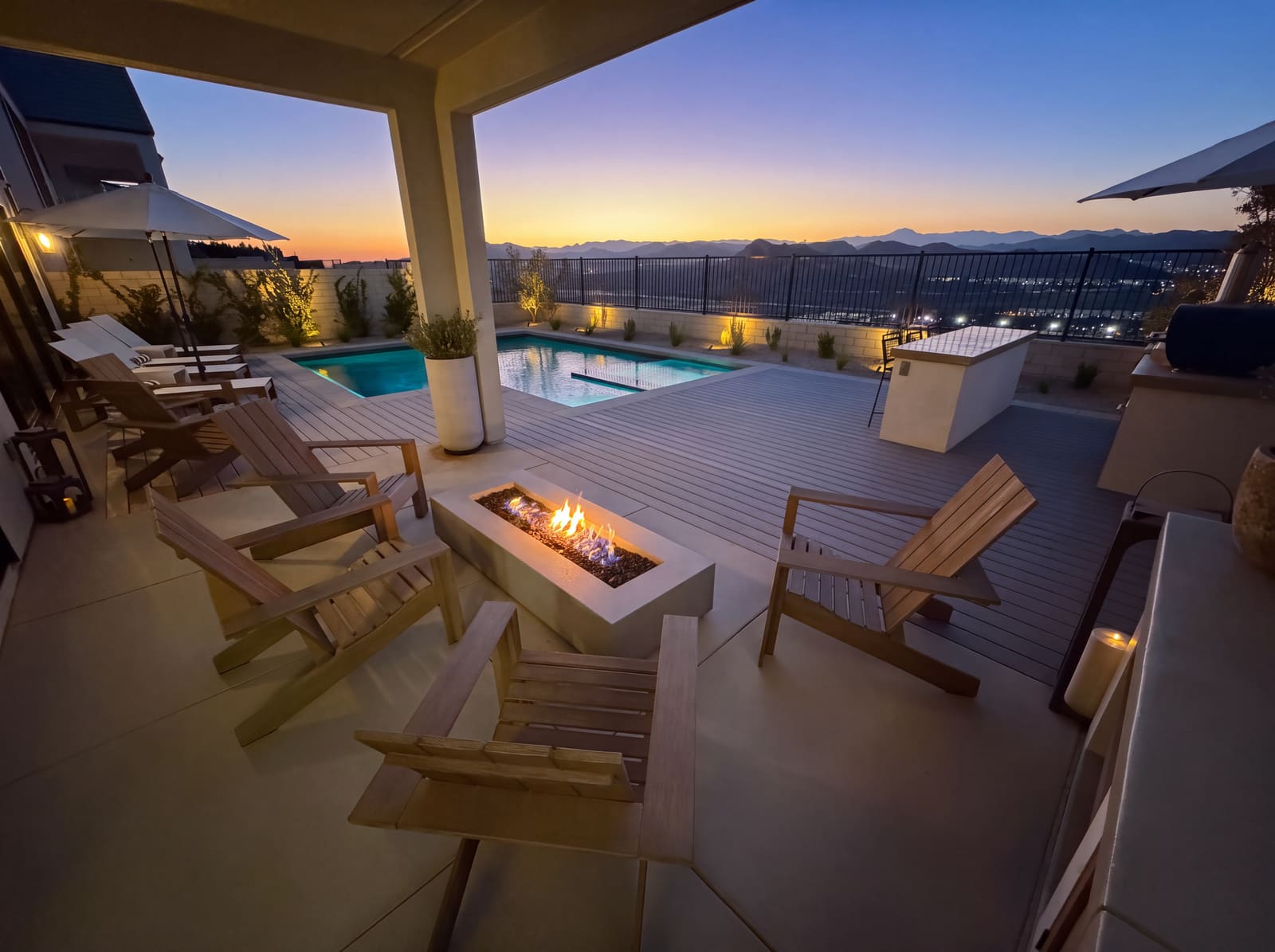 A covered deck at sunset with a lit fire table, lounge chairs, and a pool lit along its far wall above a wide valley view