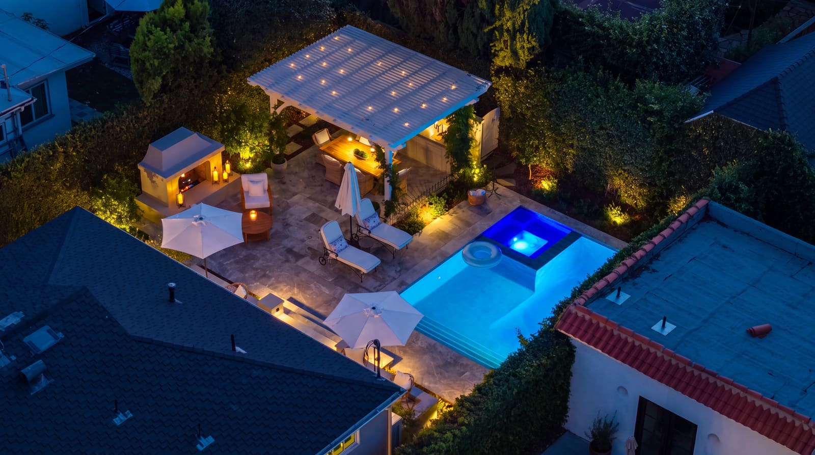 An aerial view at dusk of a lit courtyard with a pergola, an outdoor fireplace, a glowing spa and pool, and uplit planting along the walls