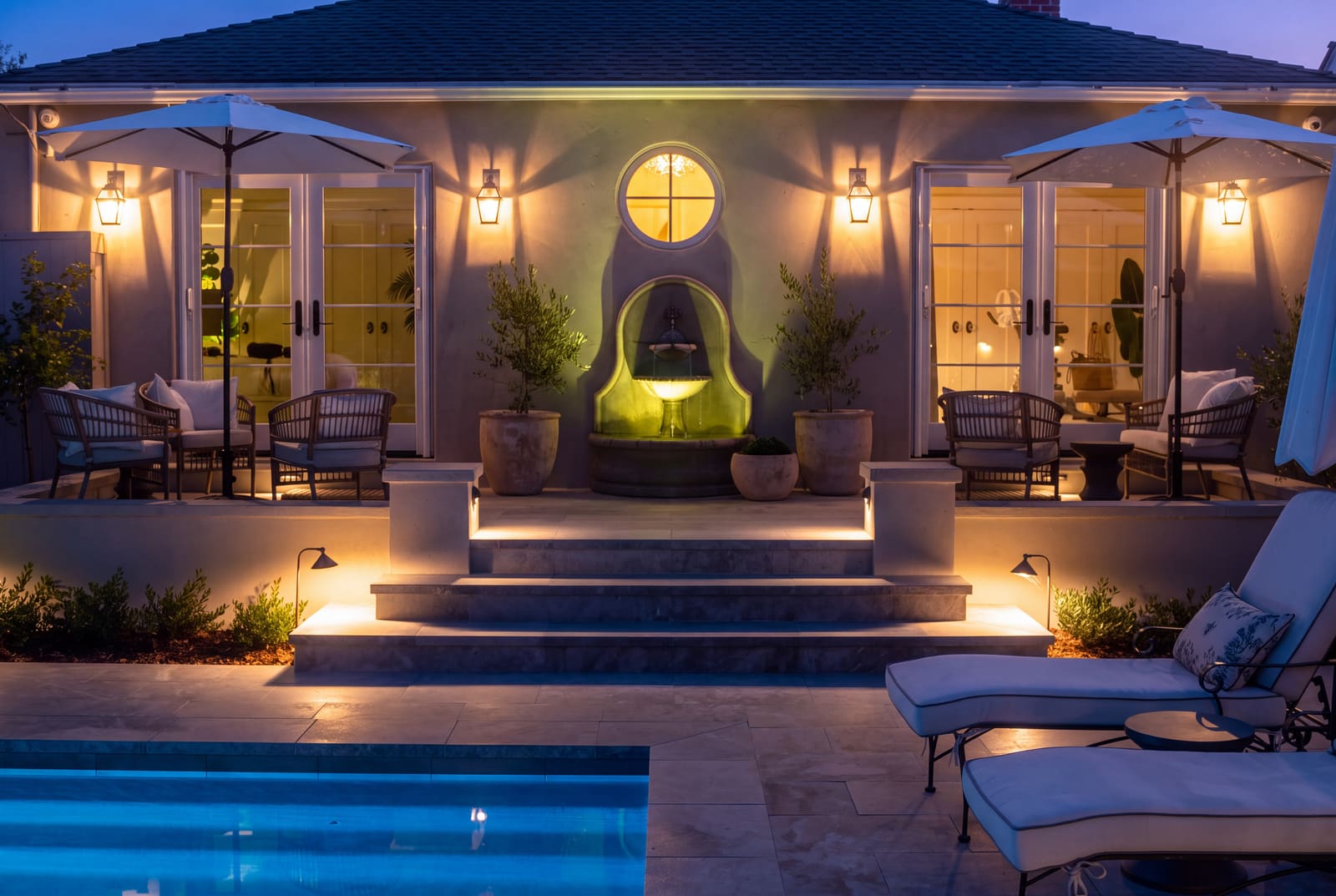 A pool terrace at dusk with lantern sconces beside two sets of French doors, an uplit wall fountain, lit steps, and path lights in the planting