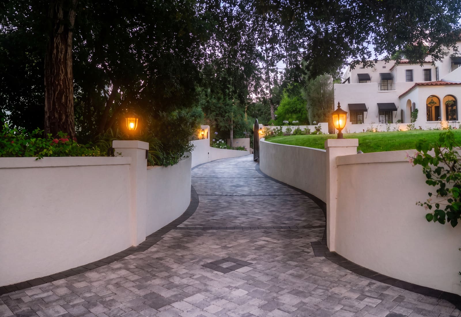 Paver driveway curving between white stucco walls, lit by lantern-topped pilasters at dusk