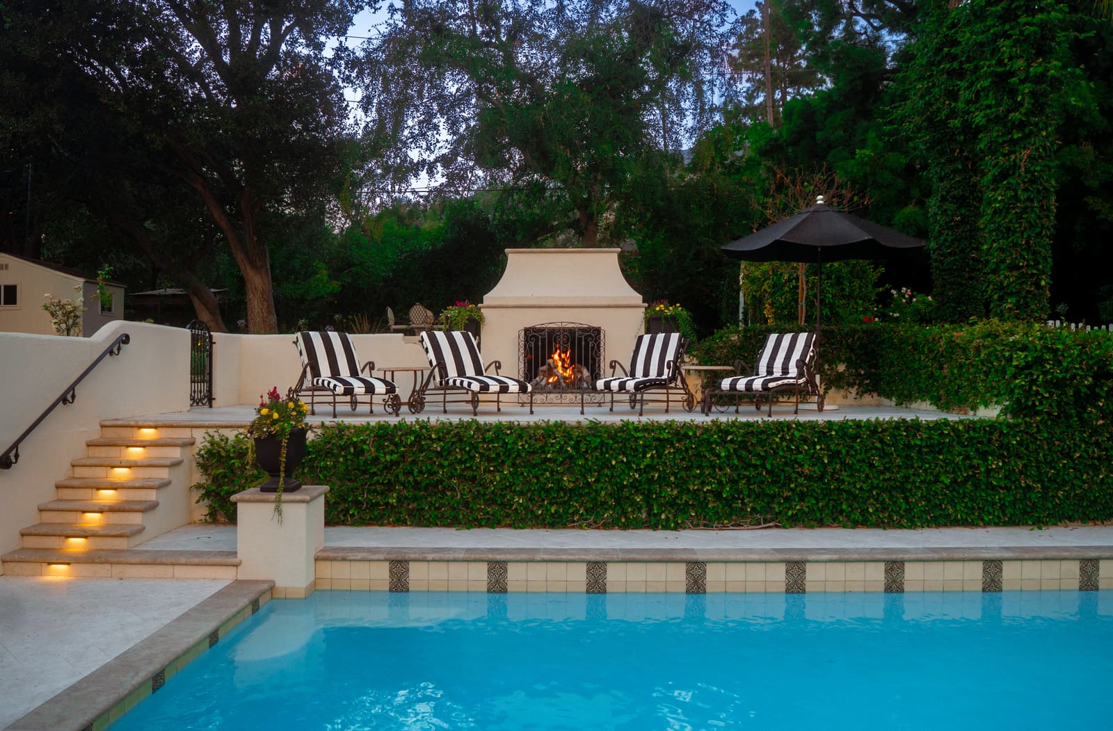 Poolside terrace with a white stucco outdoor fireplace, striped loungers and a clipped hedge running along the pool edge