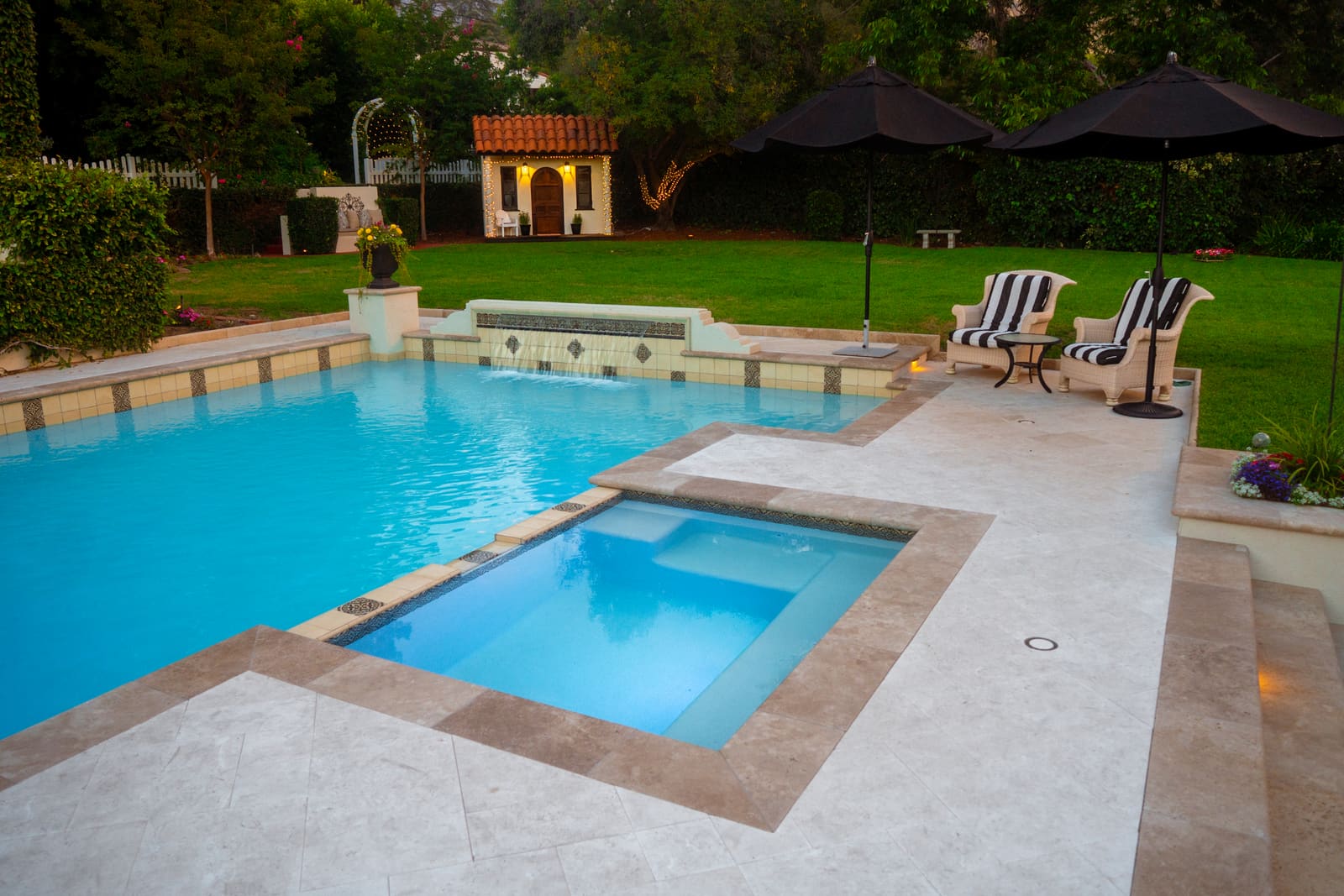 Rectangular pool and raised spa with a tiled spillway, lit from within at dusk