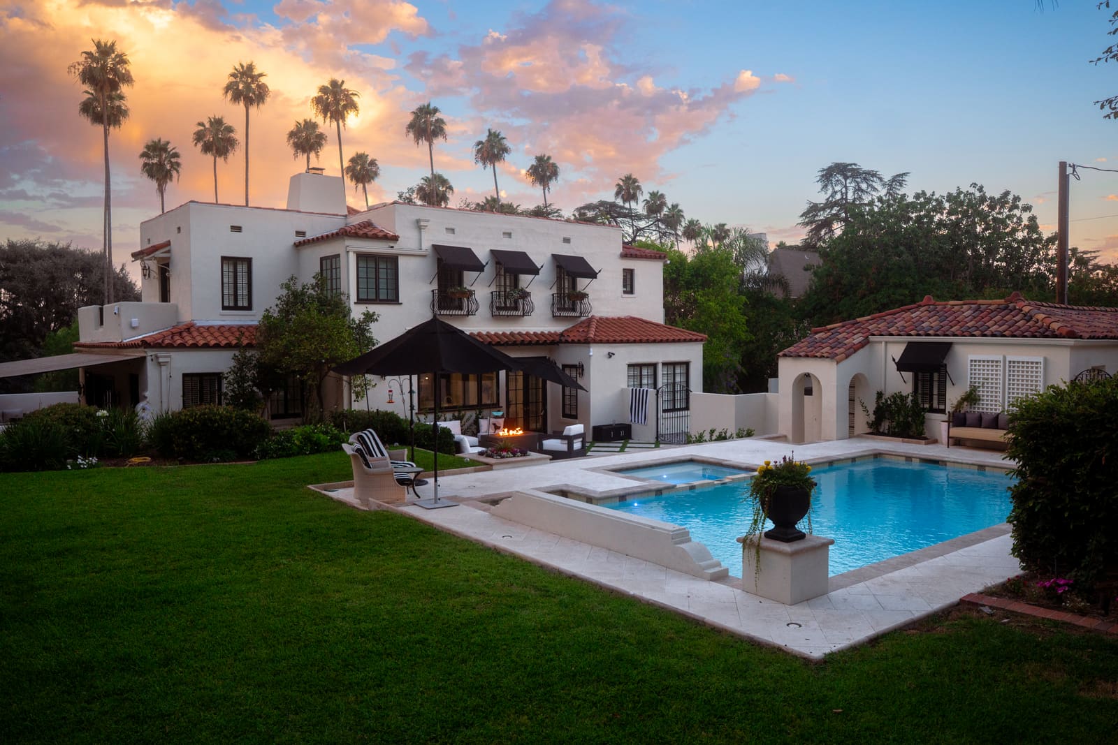 The estate's white stucco elevation and red tile roof behind the pool at sunset, framed by palms