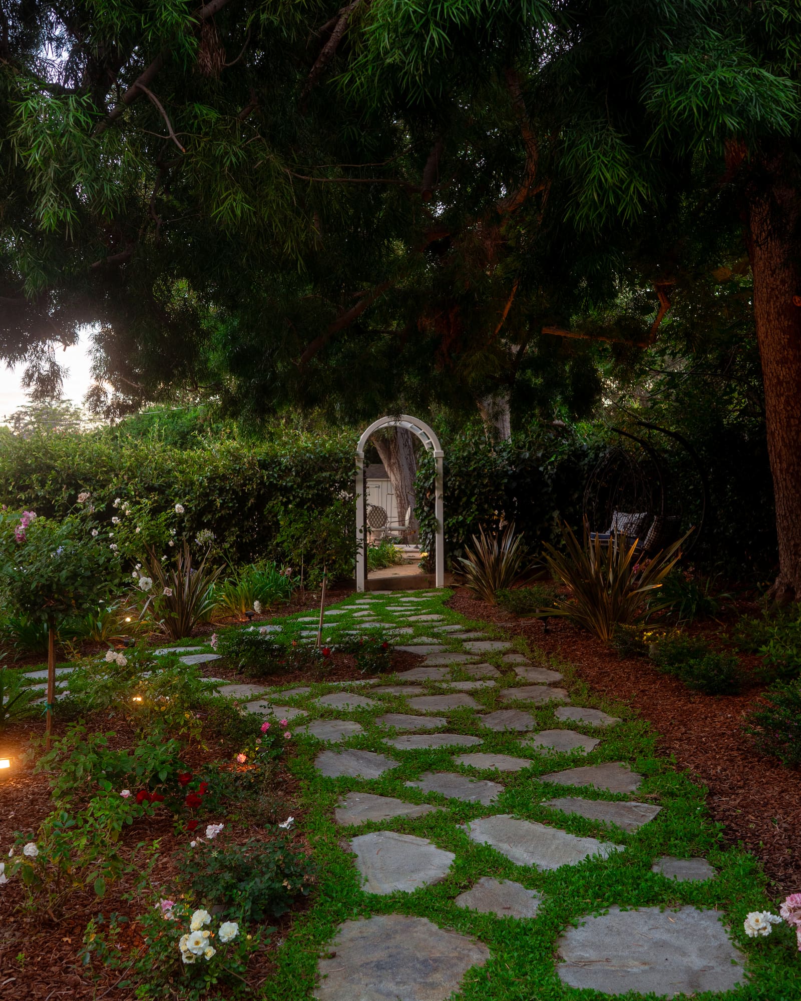 Flagstone path lit at dusk, leading through roses and flax toward a white arched gateway