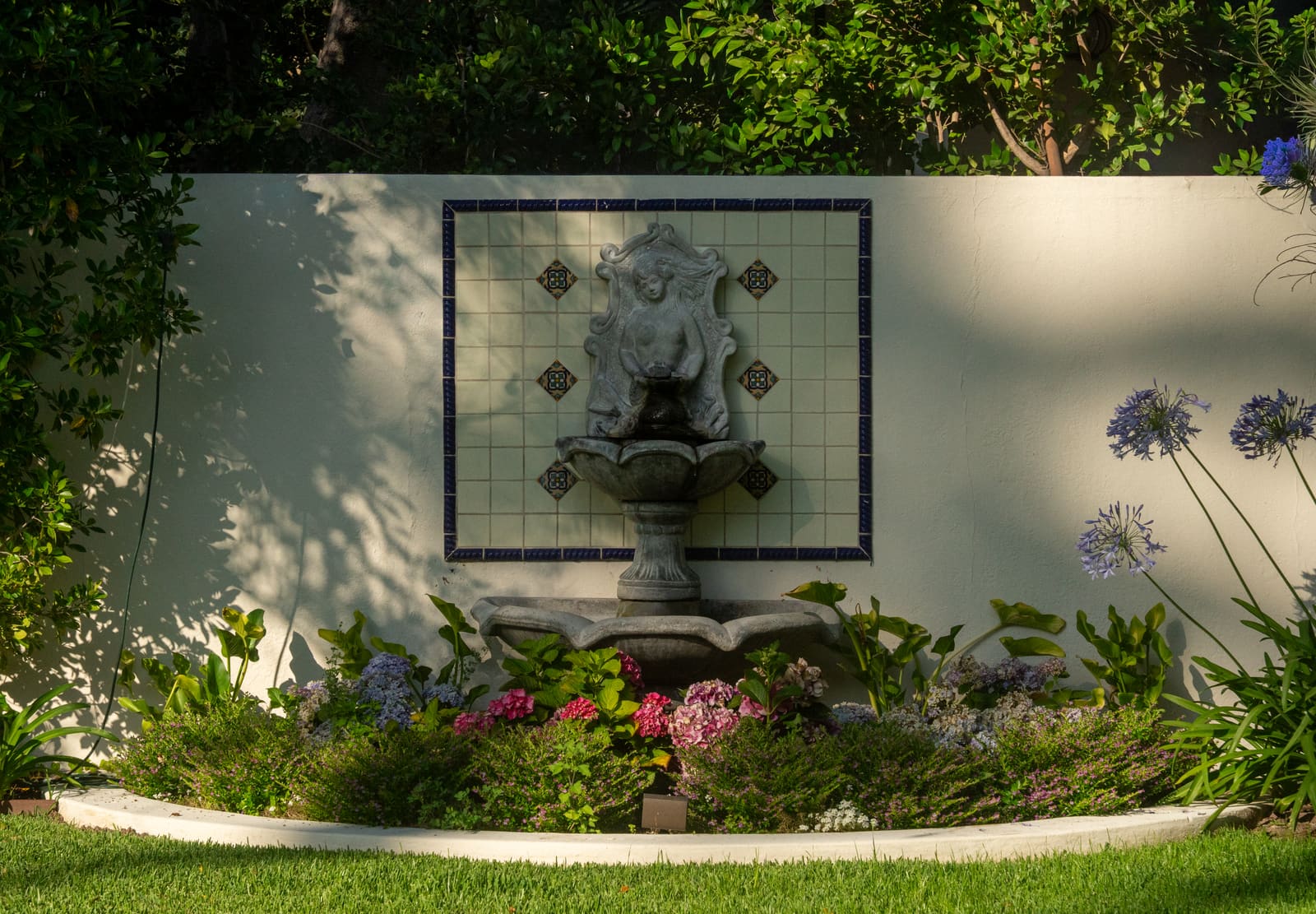 Cast stone wall fountain in a tiled surround above a bed of white alyssum