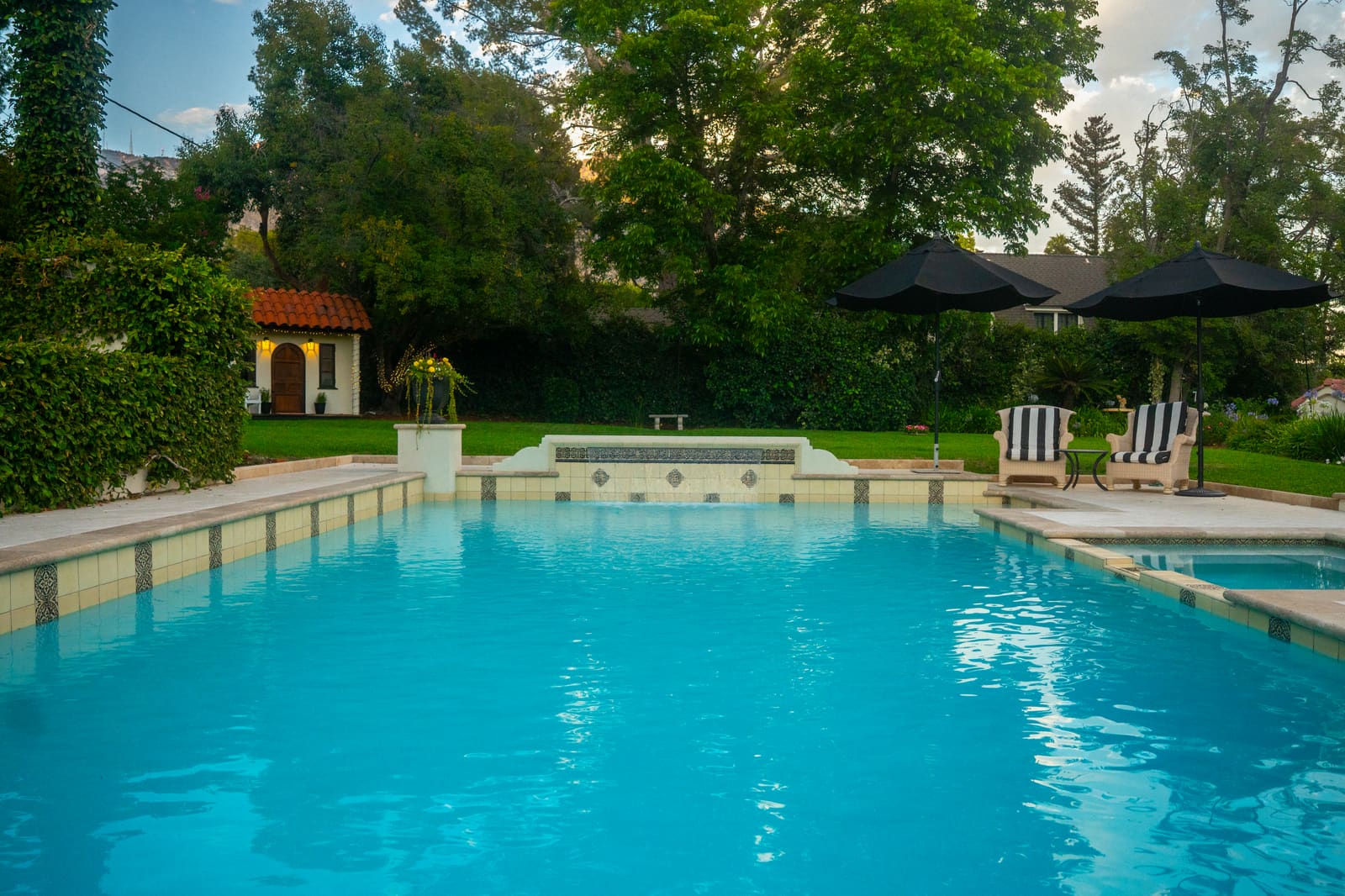 The finished pool from the deck: cream coping with a patterned tile band, a raised spillway wall, and the pool house beyond across the lawn