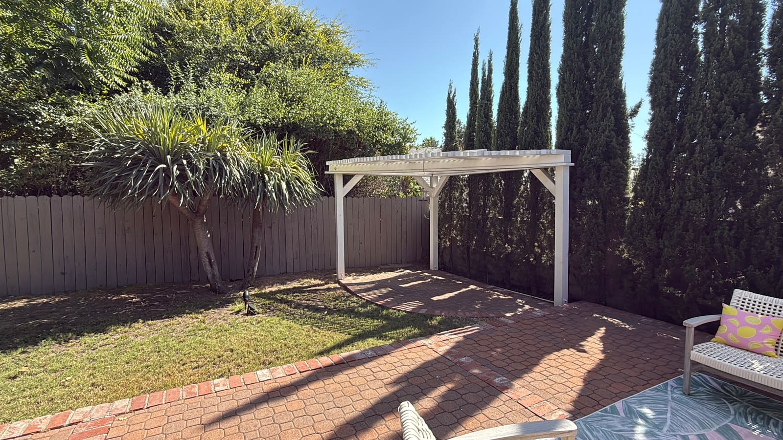 A simple existing pergola and brick patio in the backyard before construction, cypress trees along the fence