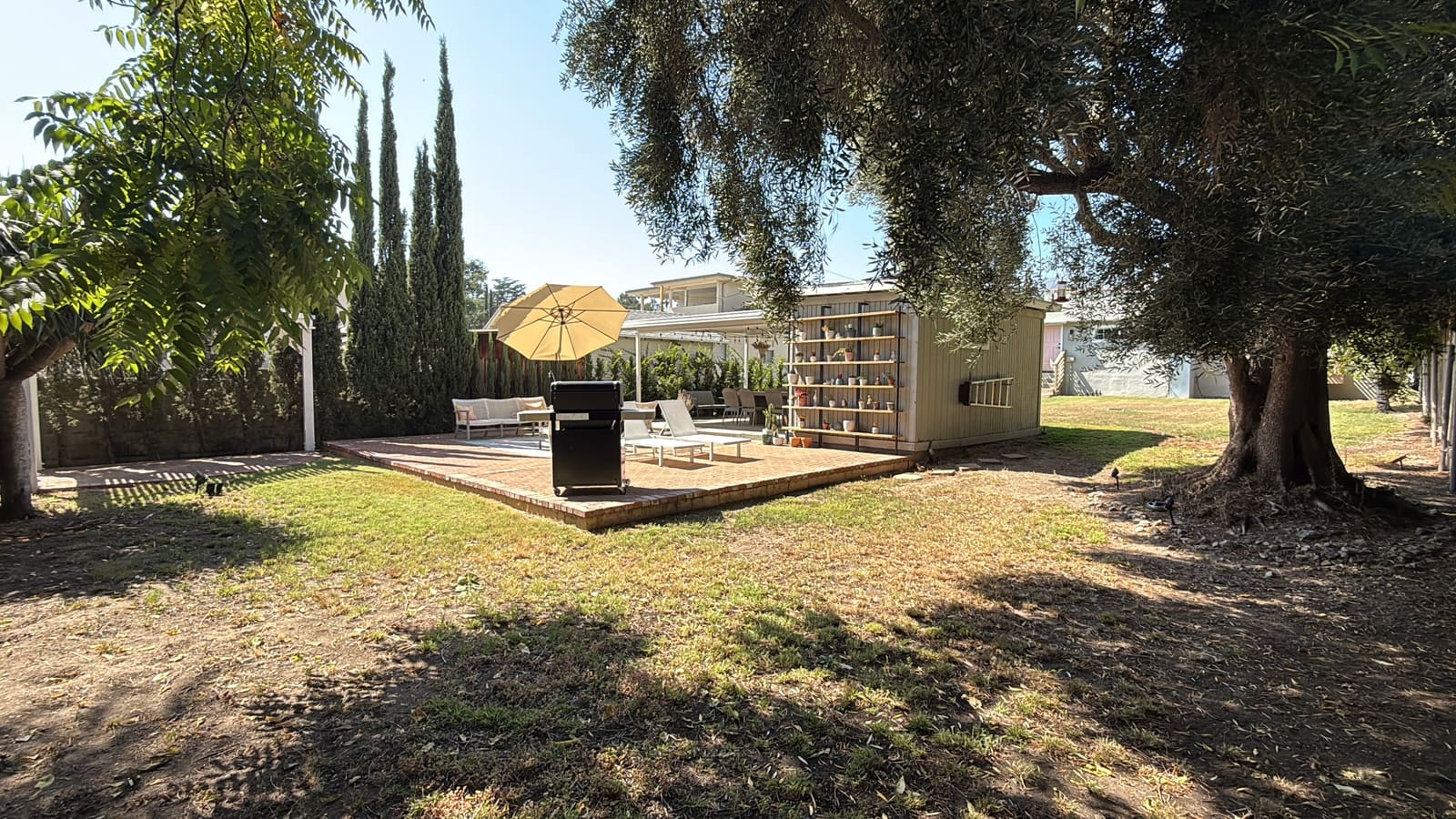 The backyard before construction, with a plain patio table, grill and storage shed on the dry lawn
