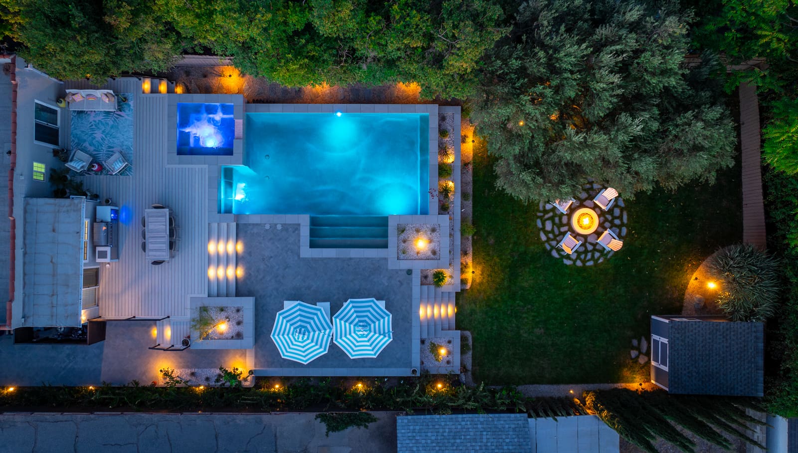 The same yard from above at dusk, with the pool, raised spa, fire pit, and step and planter lights all lit
