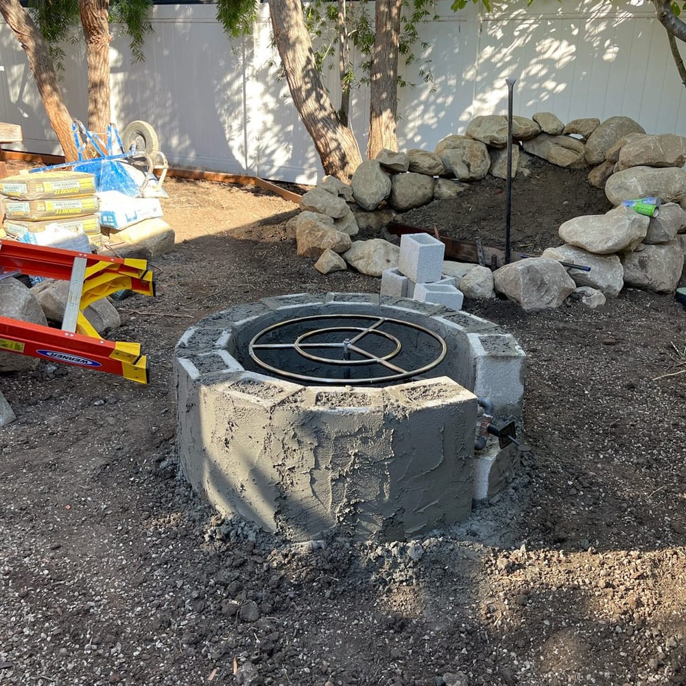 A fire pit being built during construction, with boulders already set for the water feature behind it and materials staged on site