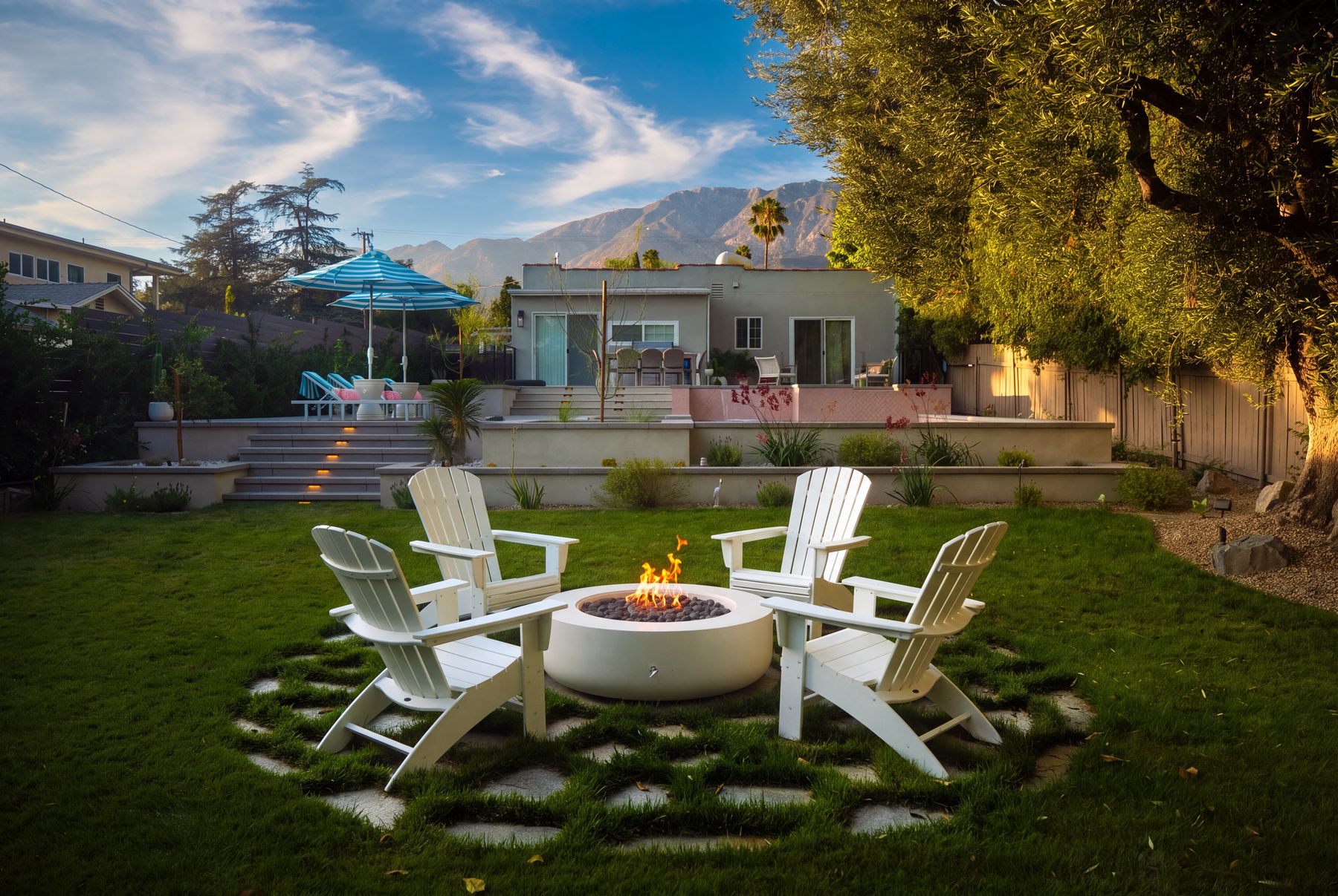 The finished fire pit lit at dusk, ringed by Adirondack chairs on a permeable paver circle with the tiered, landscaped yard behind it
