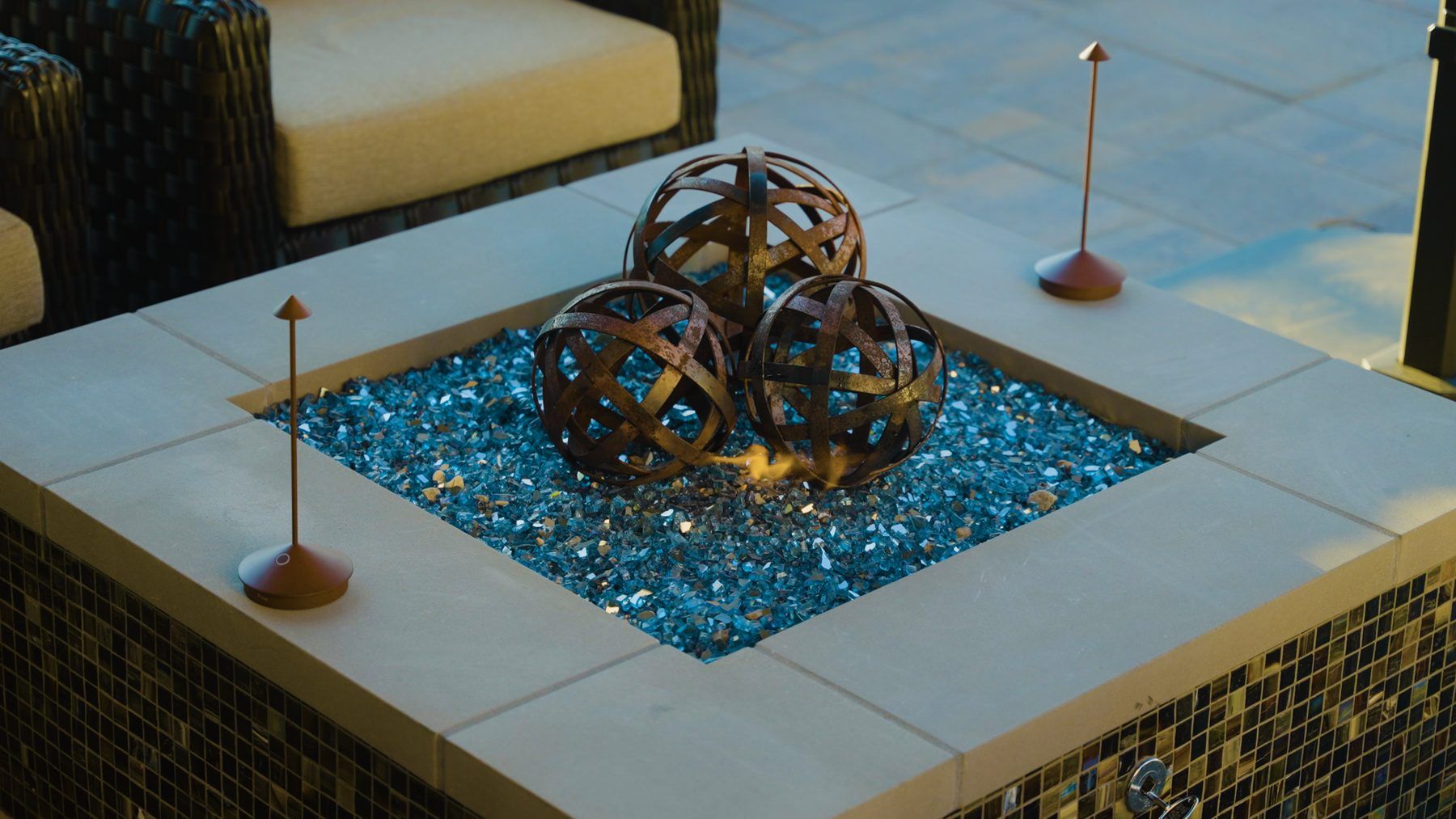 A square, tile-clad fire bowl filled with blue fire glass and a sculptural woven-metal sphere arrangement at its center