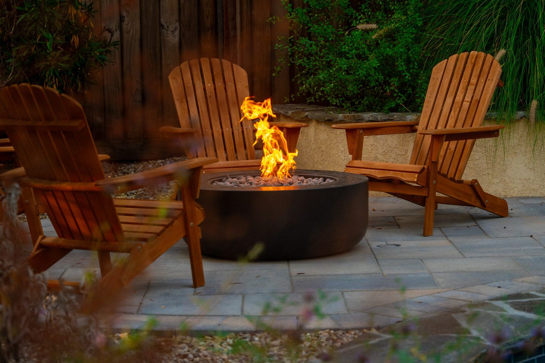 A round, free-standing fire pit ringed by wooden Adirondack chairs on a stone patio, with lava rock filling the burner