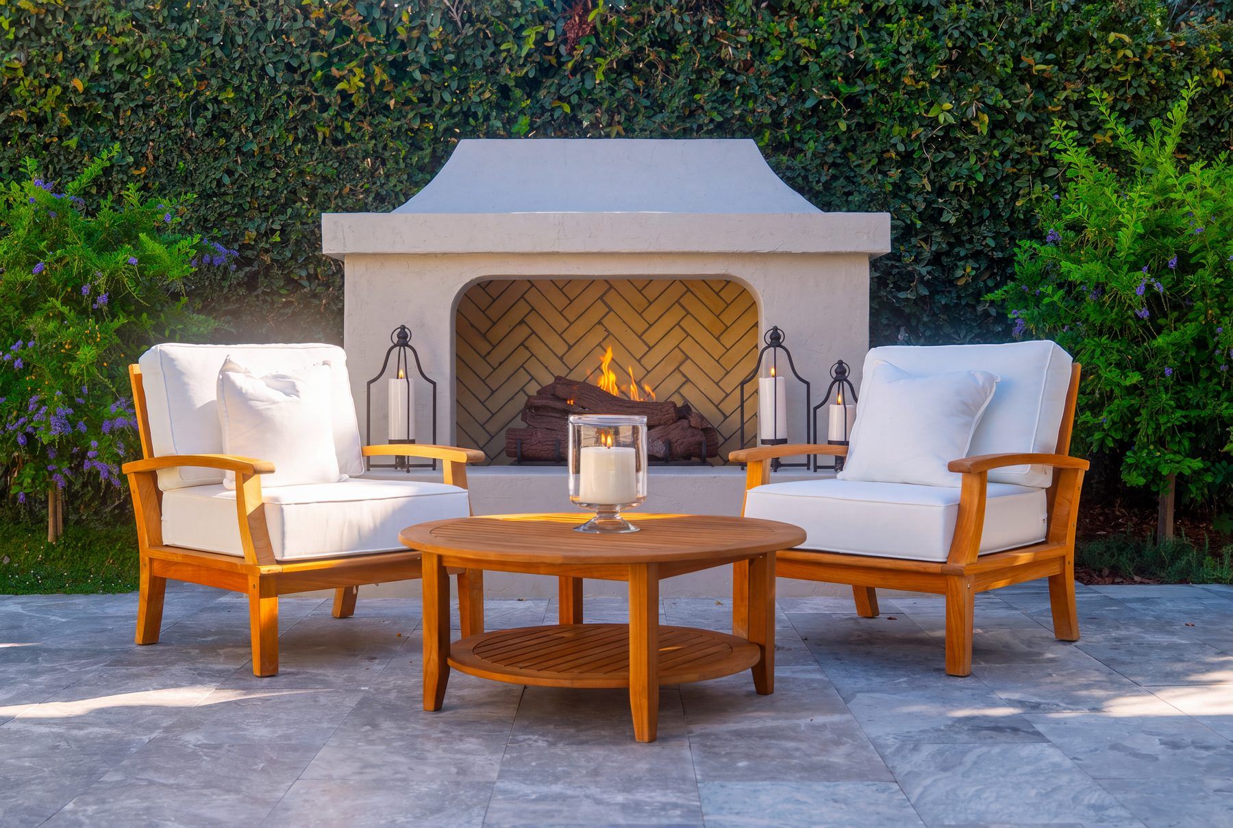 A curved stucco outdoor fireplace with a herringbone brick firebox and iron lanterns, framed by teak lounge chairs and a wisteria-covered hedge