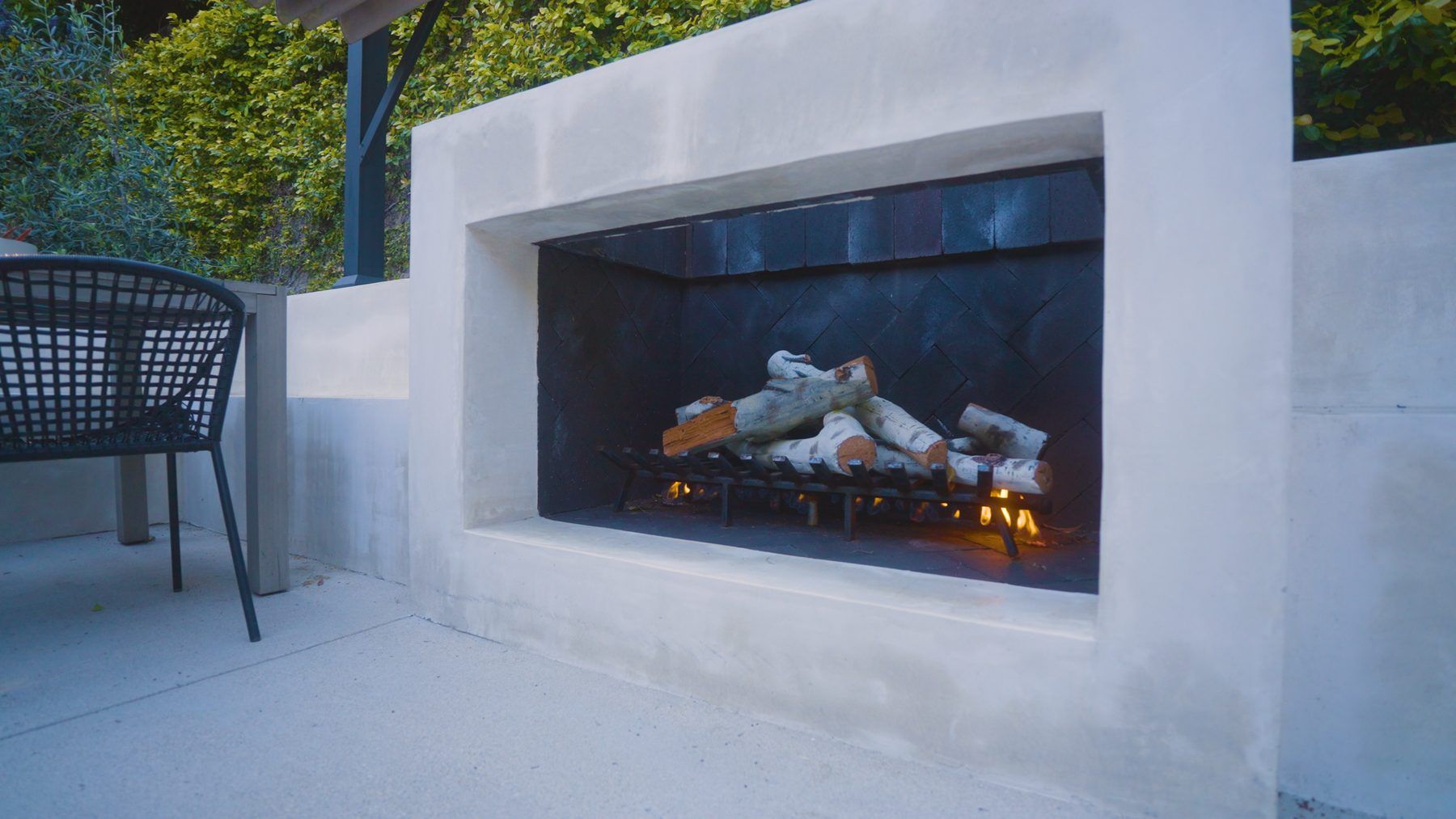 A built-in concrete outdoor fireplace with a herringbone-tile firebox interior and a lit gas log set