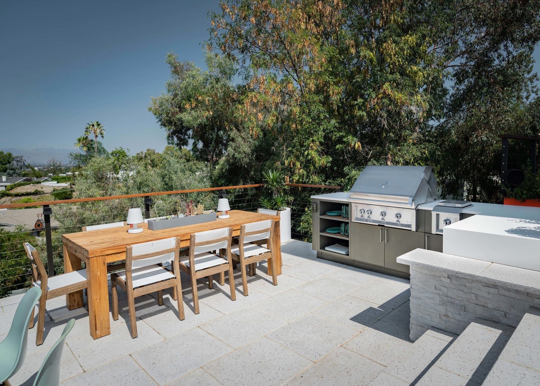 A wood dining table beside a built-in grill and counter, sharing a deck with a panoramic hillside view