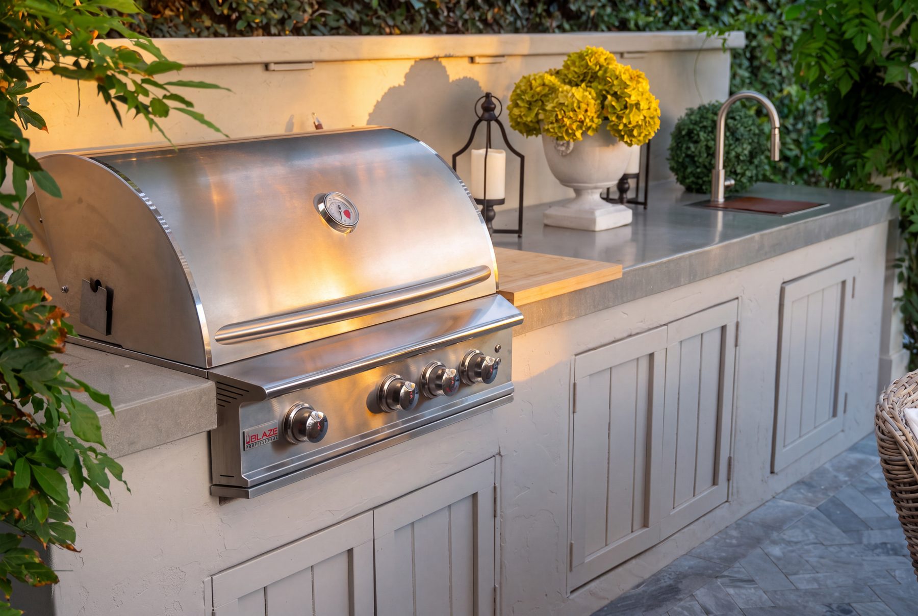 A painted Shaker-style outdoor kitchen island with a stainless grill, concrete counter, and decorative styling at dusk