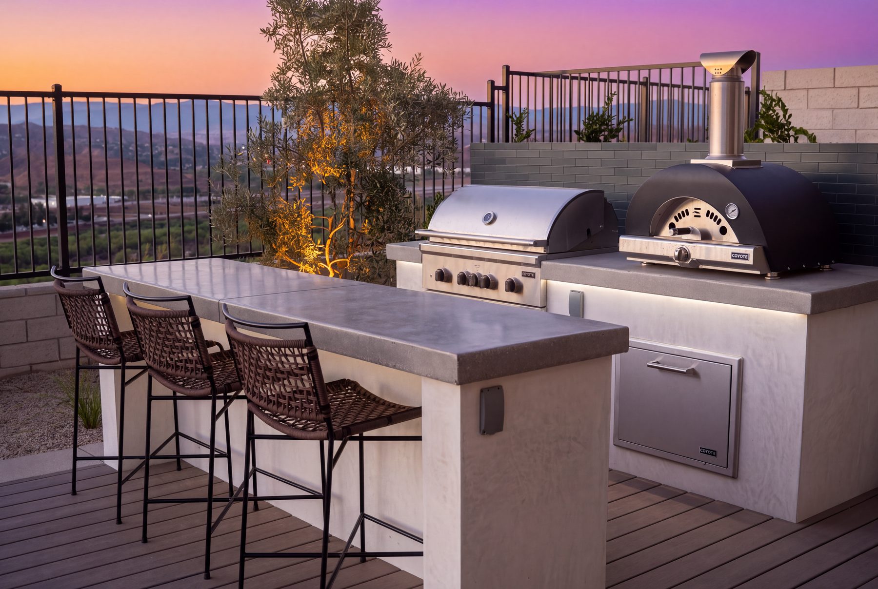 An outdoor kitchen with a grill and pizza oven beside a bar-height counter with seating, overlooking a hillside view at sunset