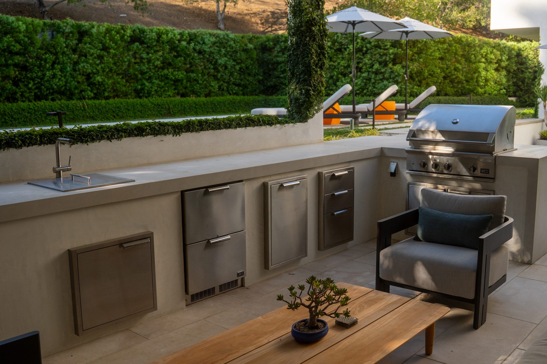 A concrete outdoor kitchen with built-in refrigerator drawers, sink, and grill, overlooking a pool and hillside