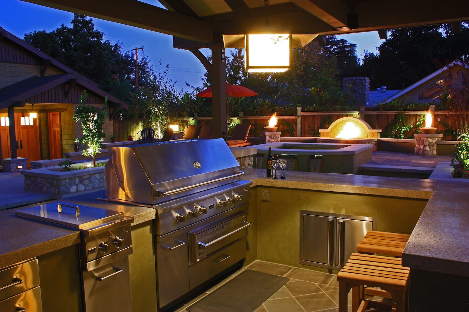 A craftsman patio at dusk with a stainless grill beside a tall stone fire pit and two lit fire bowls, string lights overhead