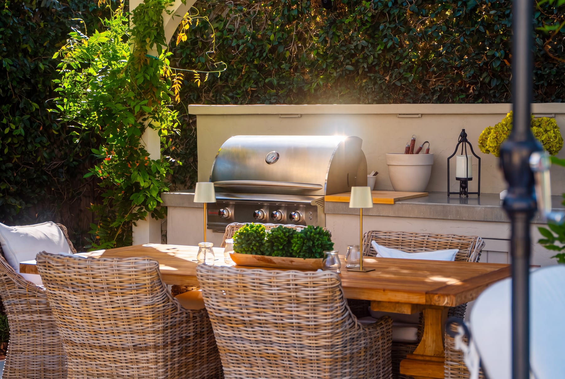 A stainless grill and prep counter under a flowering vine-covered trellis, with a lantern and potted greenery at golden hour