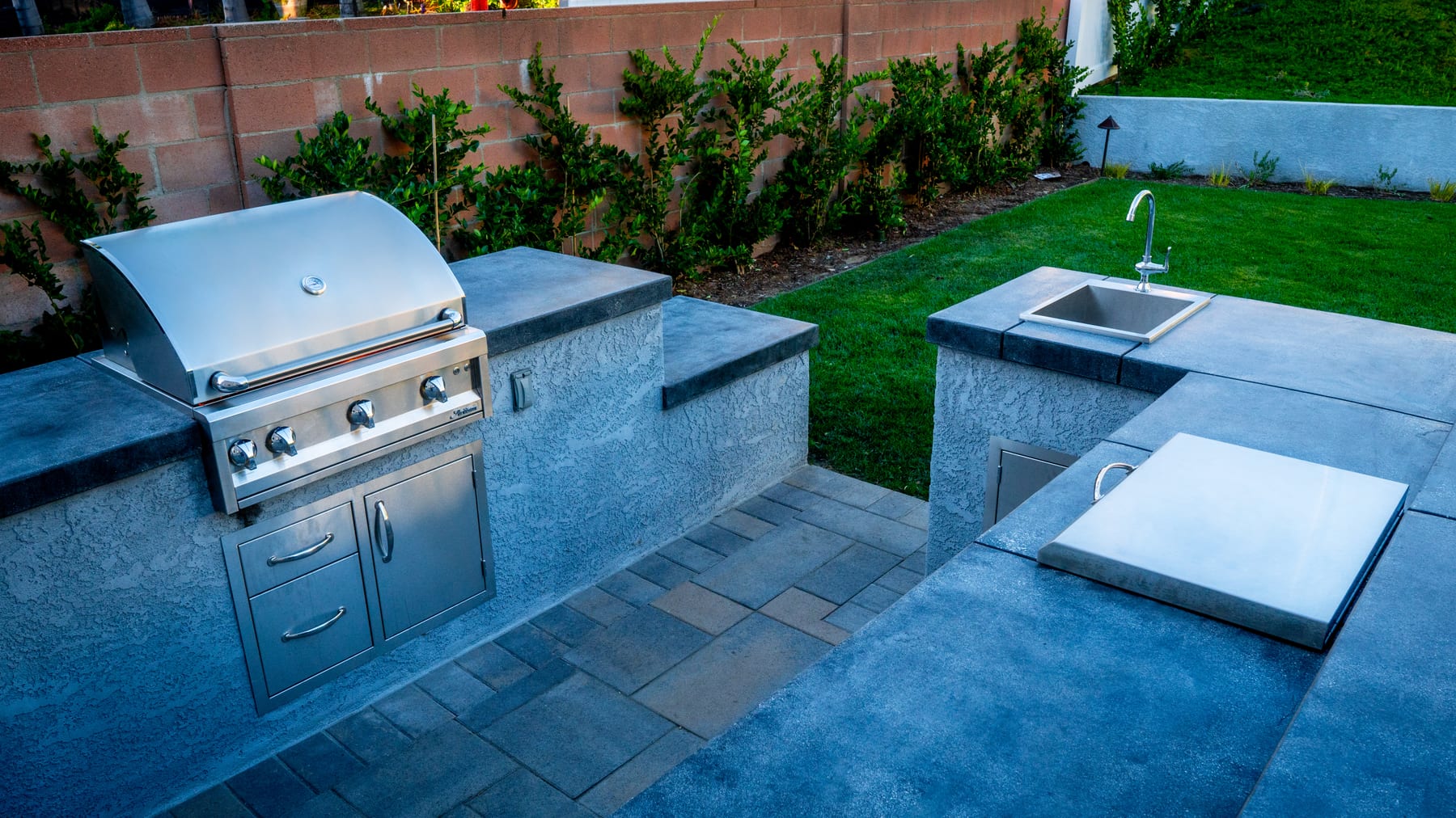 A stone-clad outdoor kitchen with a stainless grill and built-in sink beside a manicured lawn at dusk