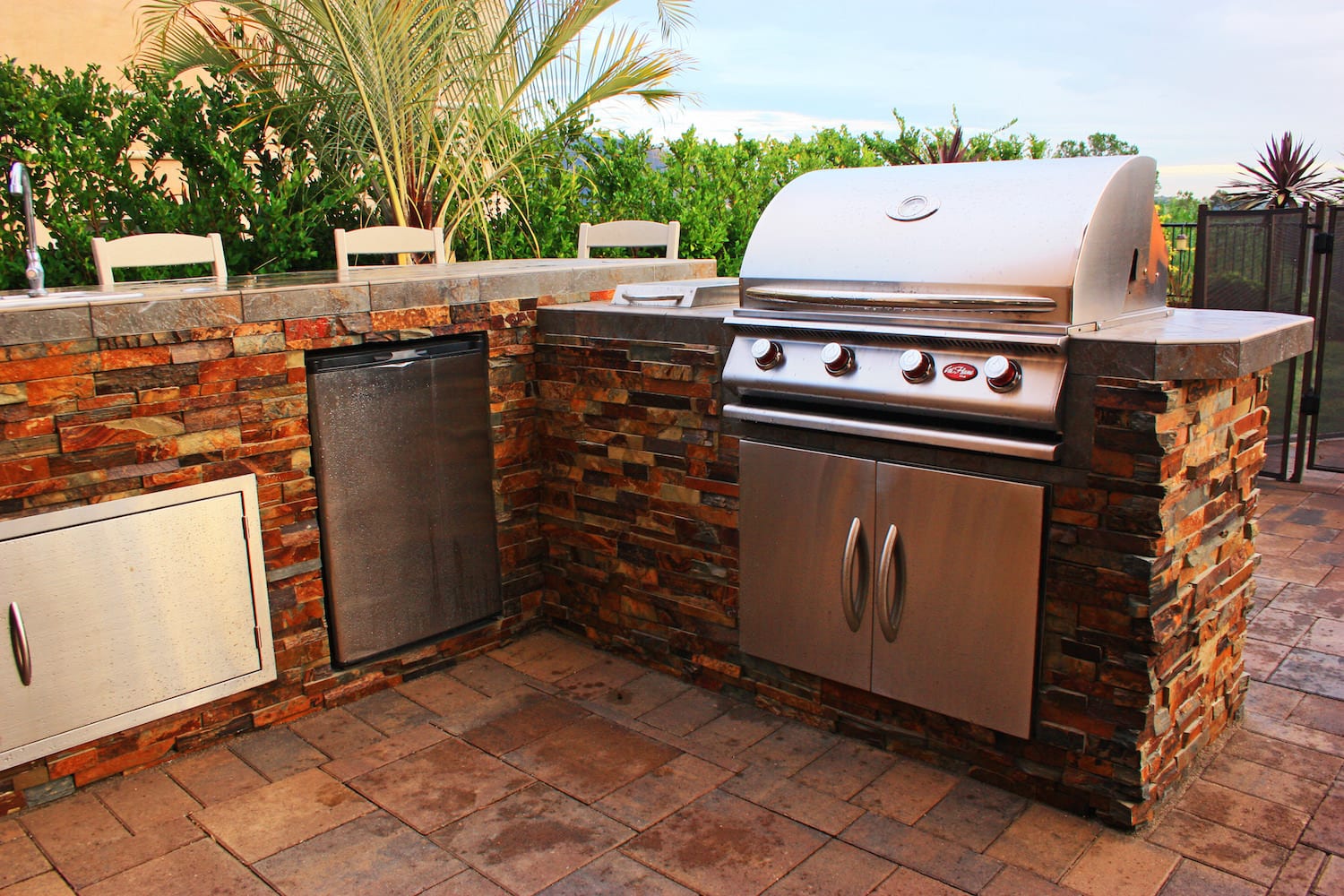 A multicolor stone-clad outdoor kitchen island with a stainless grill and built-in fridge on a paver patio at dusk