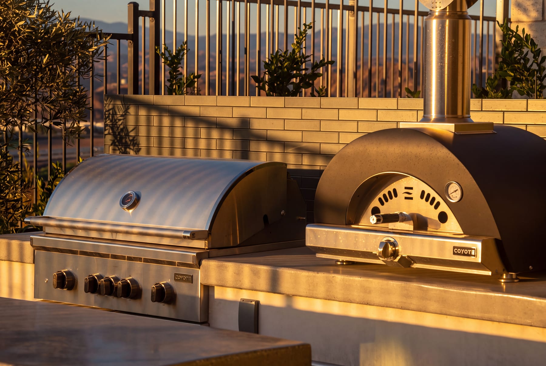 A rooftop outdoor kitchen with a stainless grill and wood-fired pizza oven overlooking distant hills at golden hour