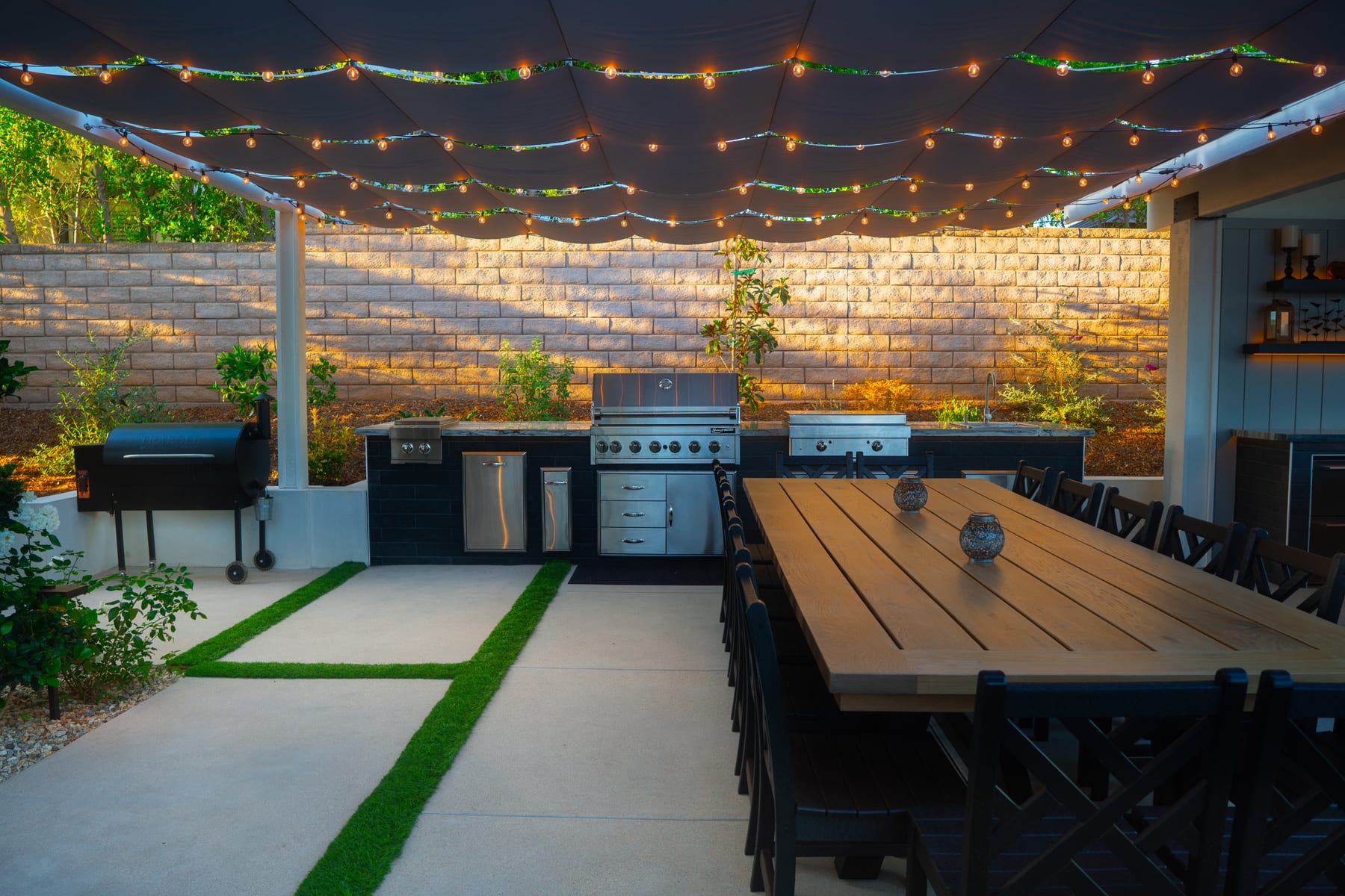 An outdoor kitchen island with a smoker, grill, side burner, refrigerator drawers, and sink under a string-lit canopy at dusk