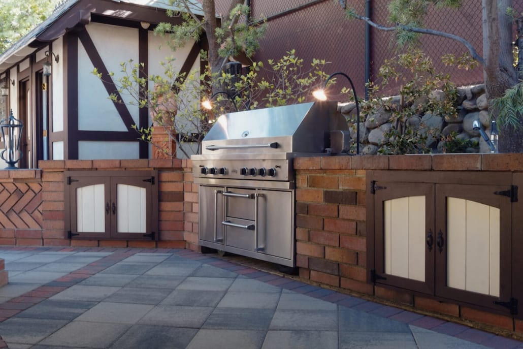 A brick outdoor kitchen with a stainless grill and white cabinet doors built into a low wall, backed by a stacked river-rock retaining wall