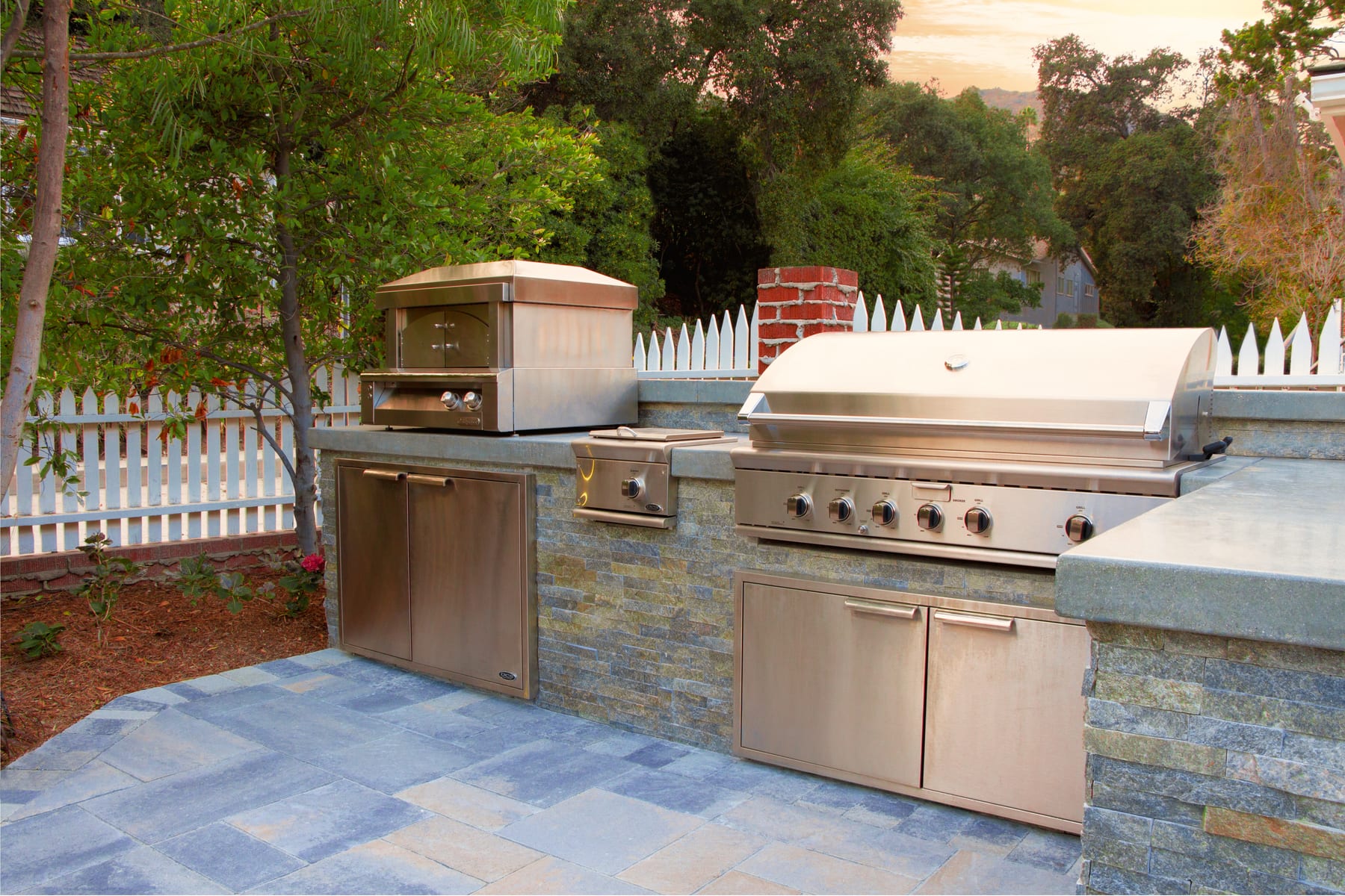 A stainless pizza oven and grill on a stone counter at dusk, framed by a white picket fence and a brick chimney pillar