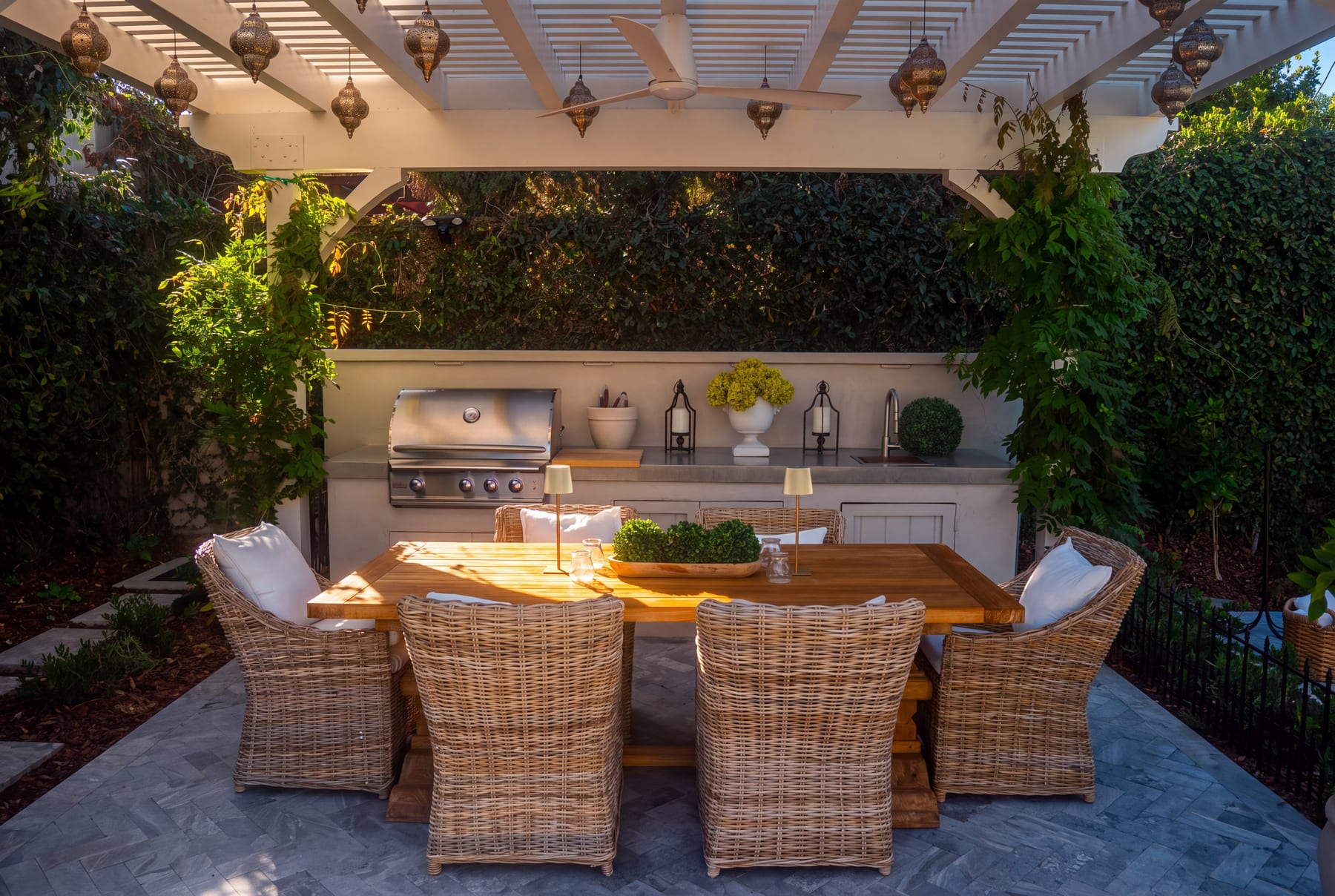 A wisteria-draped pergola with hanging lanterns over a long dining table and wicker armchairs beside a built-in grill