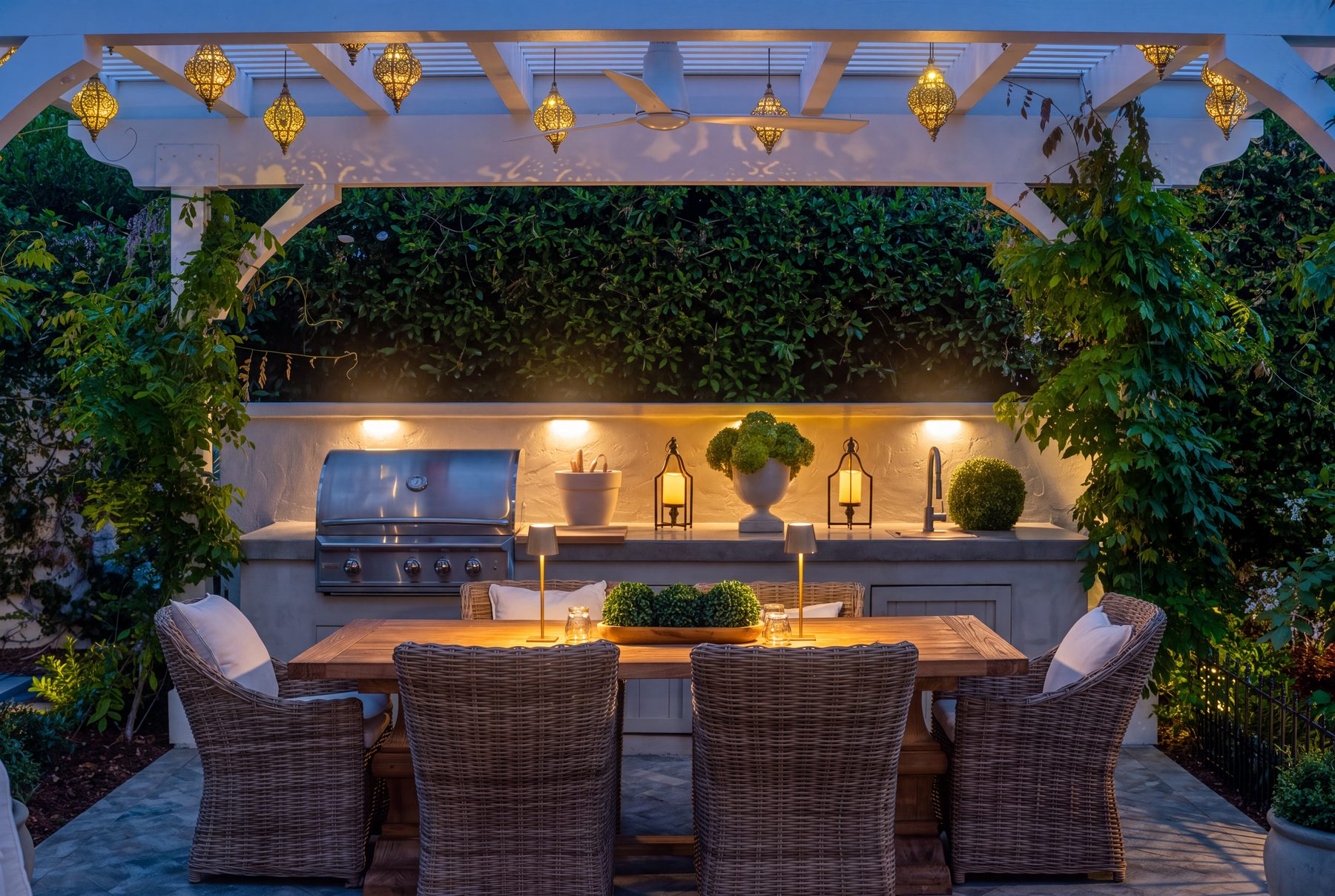A wisteria-covered pergola strung with Moroccan lanterns over a built-in grill, stone counter, and wicker dining set at dusk