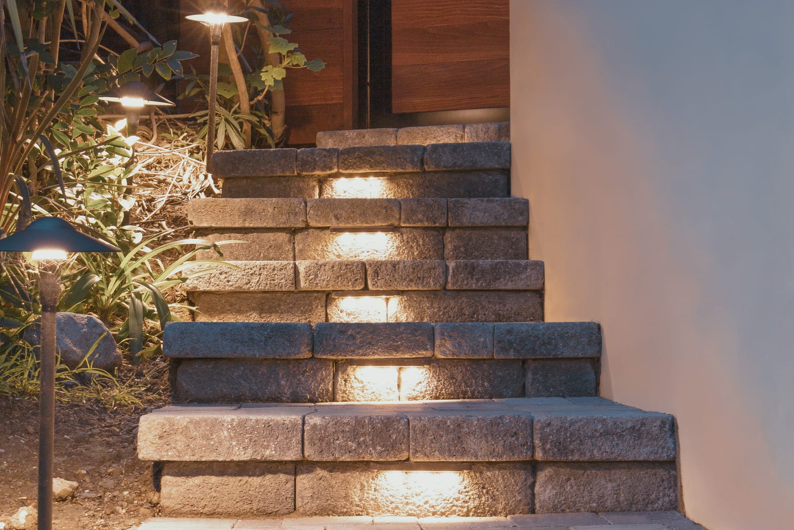 Concrete block steps lit by step lights in each riser, with downward-facing path lights in the planting beside a wood gate at night