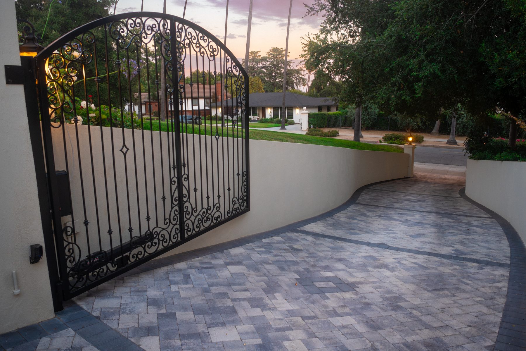 A wrought-iron gate opening onto a paver driveway at dusk, with a single-story home visible beyond