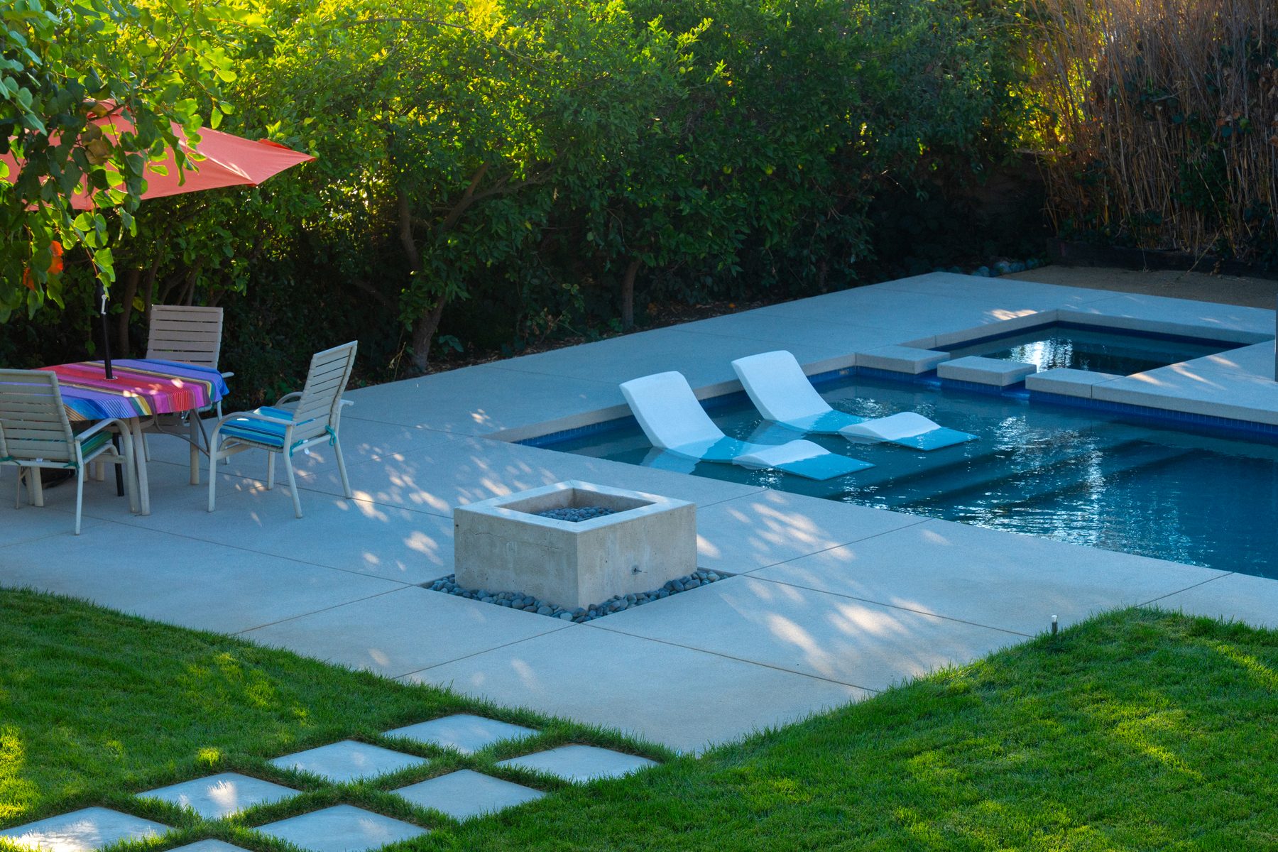 A pool deck patio with a built-in square fire pit, a striped-cloth dining table, and in-water lounge chairs