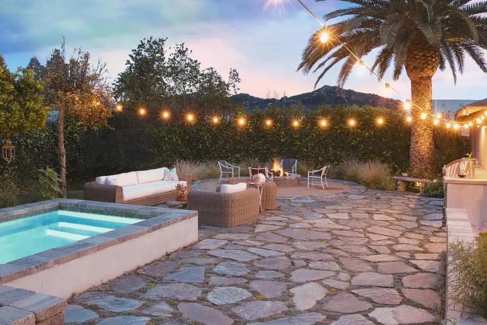 A flagstone patio at dusk with a raised spa, string lights, a fire pit lounge area, and a hillside view
