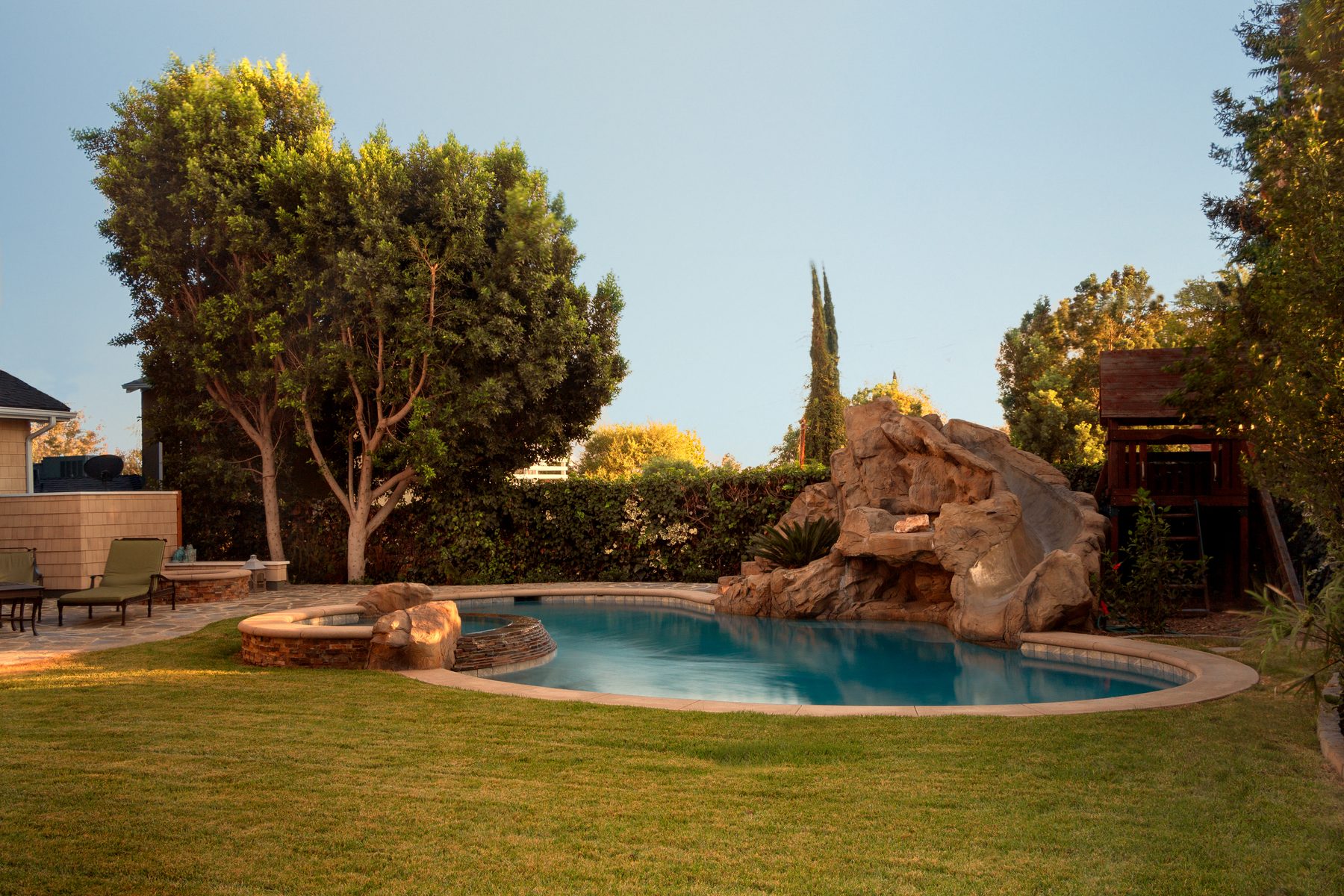 A freeform pool with a boulder waterslide and spa, a flagstone patio, and a lawn with a play structure