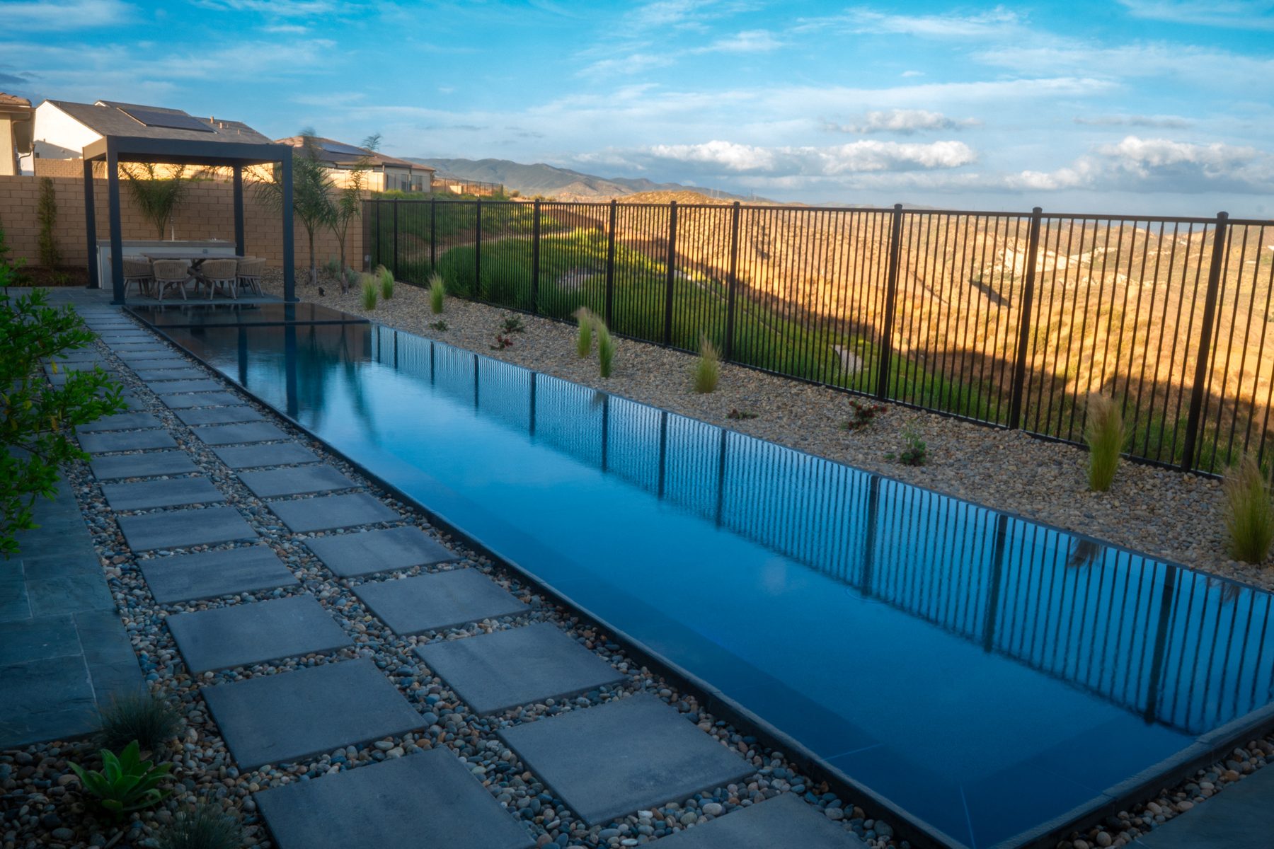 A hillside infinity-edge lap pool with a covered dining pergola and rolling rural view beyond an iron fence